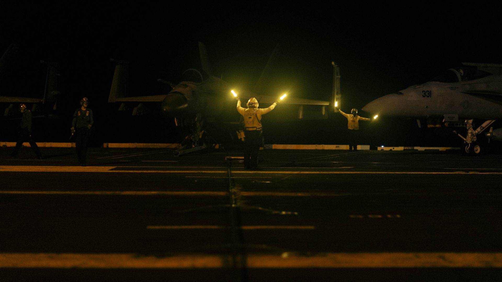 Nighttime scene on aircraft carrier deck with crew in yellow jackets using illuminated wands to guide two stationed fighter jets.