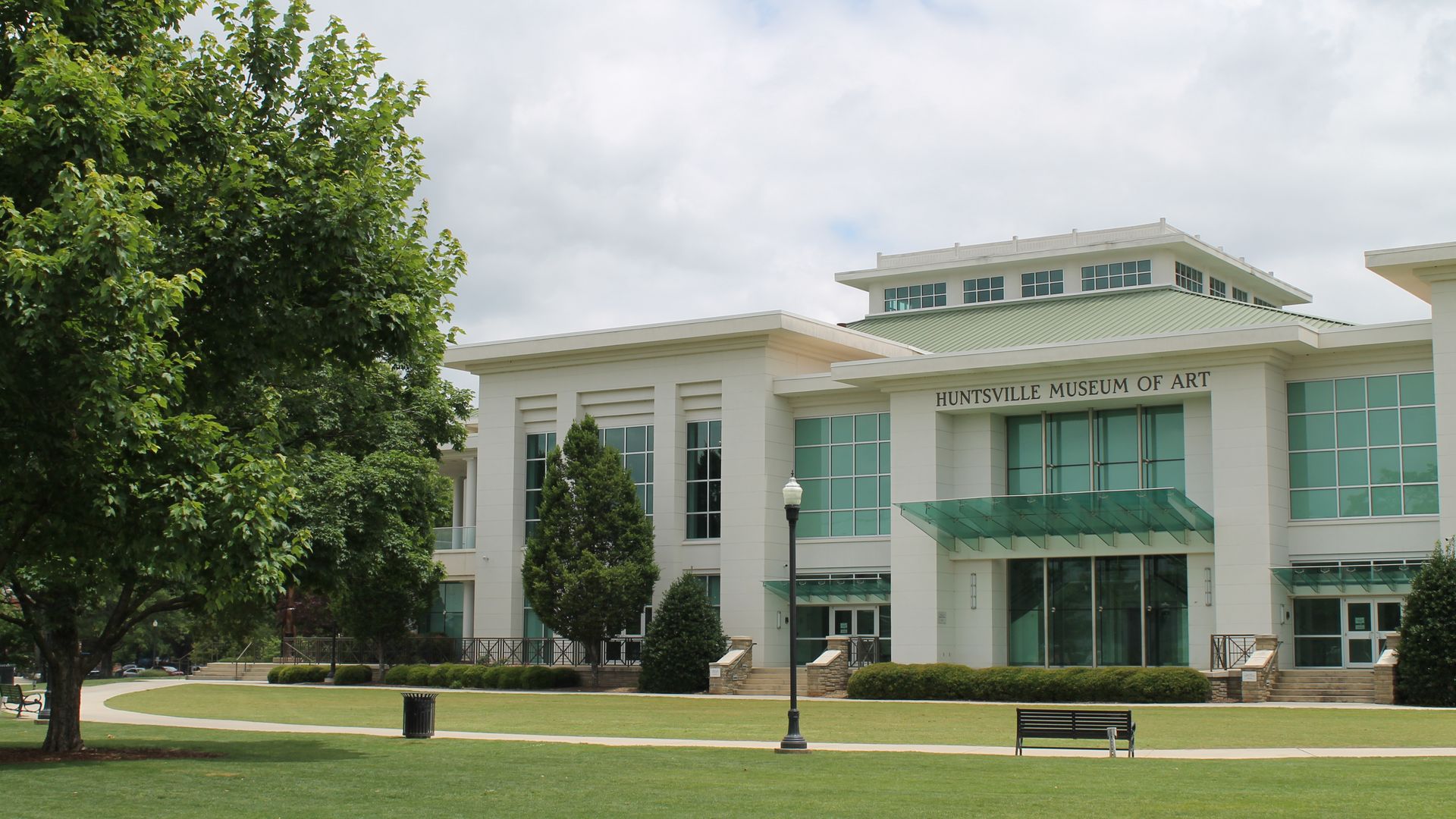 Modern Huntsville Museum of Art building with green roof and large windows, surrounded by green grass, trees, a black bench, and a lamppost under a cloudy sky.