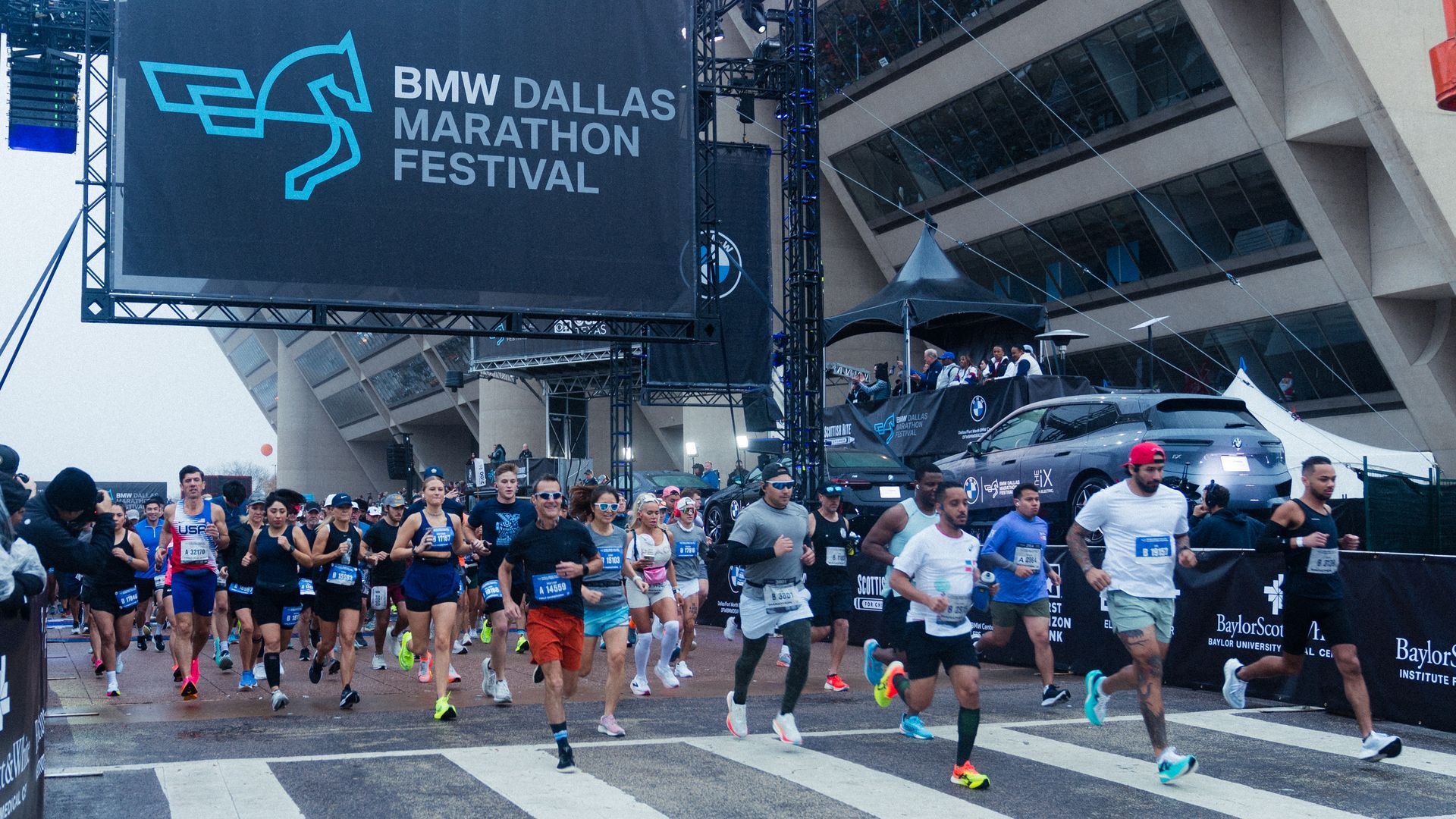 Runners start the BMW Dallas Marathon Festival race, crossing a marked street under a large black sign with a blue horse logo and race title, with spectators and event staff nearby.