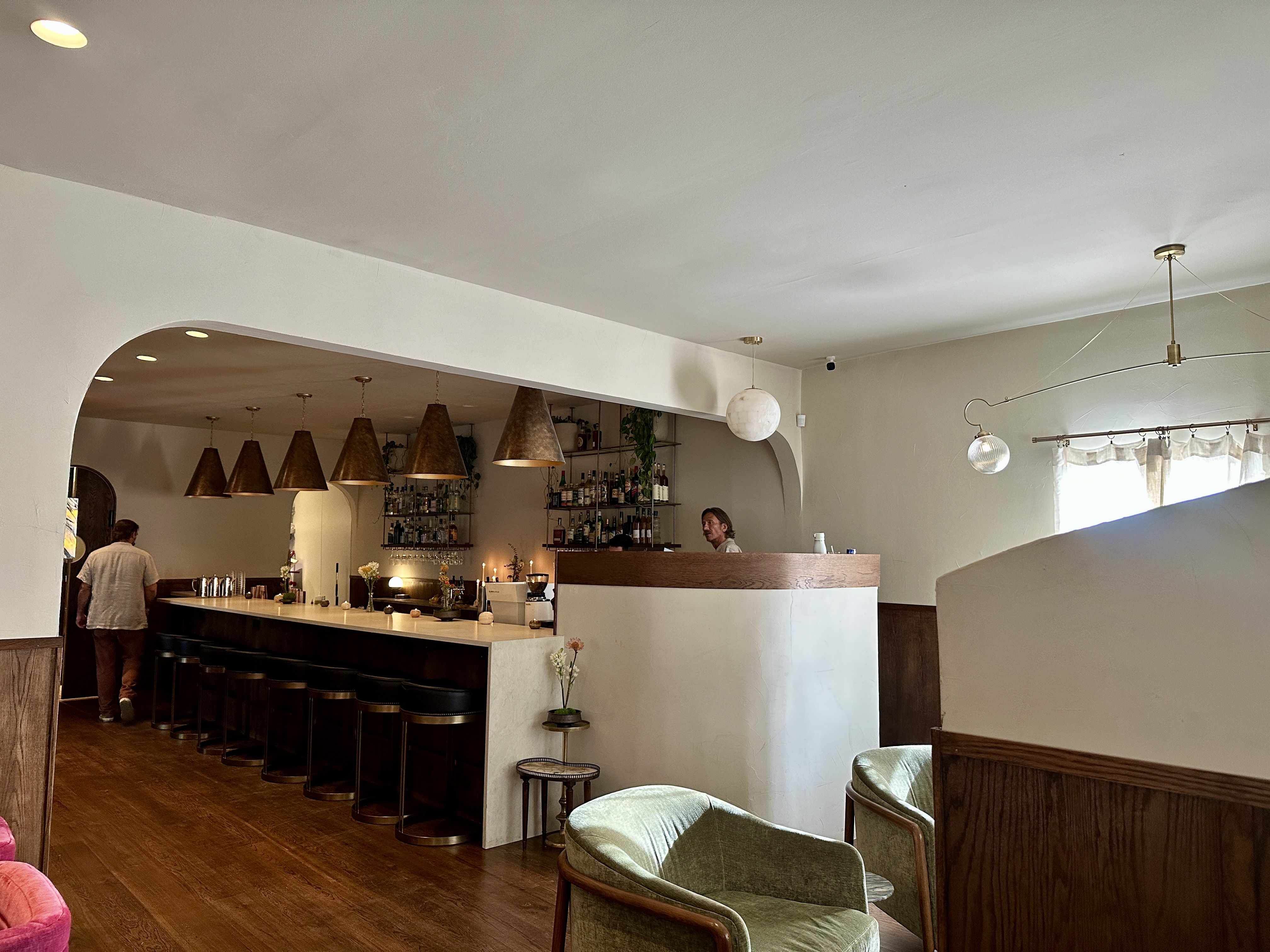 Interior of a modern cafe with white curved walls, a rounded arch opening, and a long wooden bar with brown stools. Bottle shelves, warm pendant lights, and globe lamps. Huckleberry Noda now open.