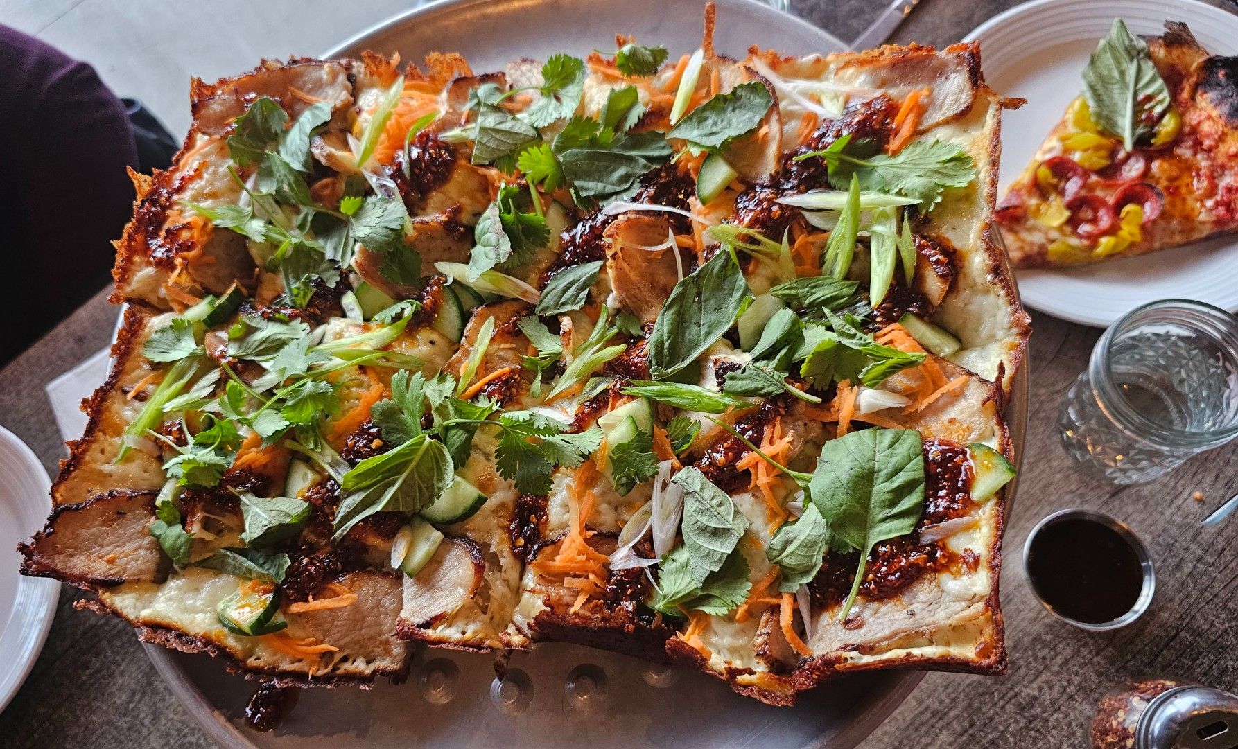 Overhead view of a large rectangular flatbread/pizza topped with fresh cilantro, mint, cucumber, shredded carrots and green onions; a dark sauce drips over the crispy crust, served with a side dish and a water glass.