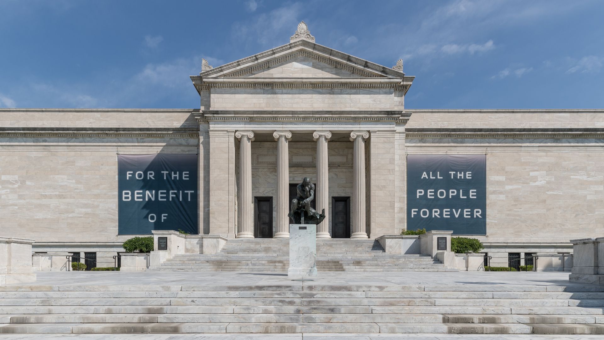 Neoclassical marble museum with six tall columns and a triangular pediment; statue at the stairs; blue banners read "FOR THE BENEFIT OF ALL THE PEOPLE FOREVER" against a clear blue sky.