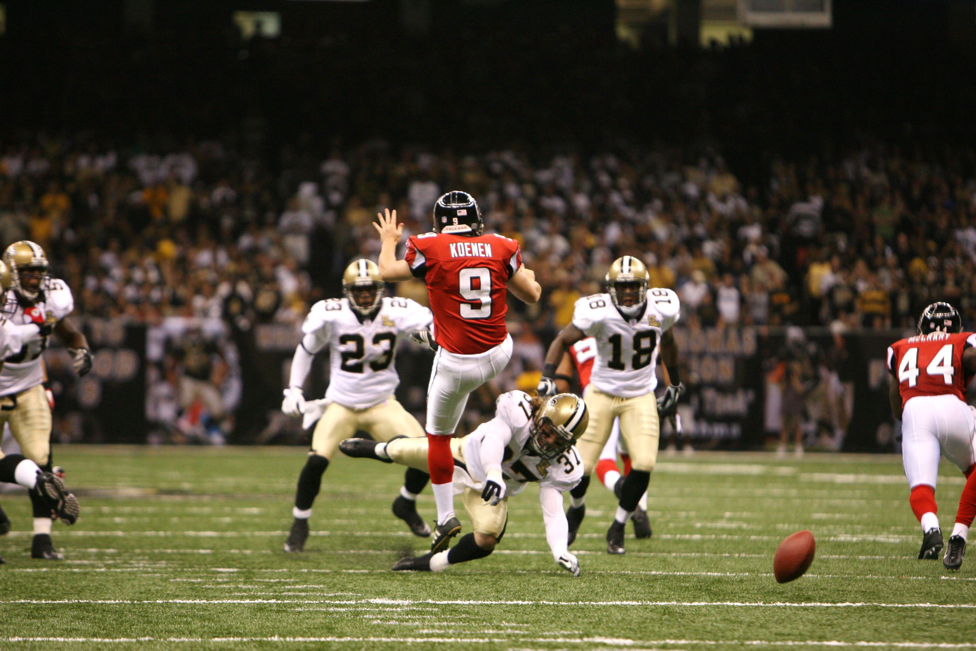 Action during a football game at the Superdome. A player wears No. 9 in red, immediately after kicking. His arm is raised while the football bobbles off to the right. Saints players approach.