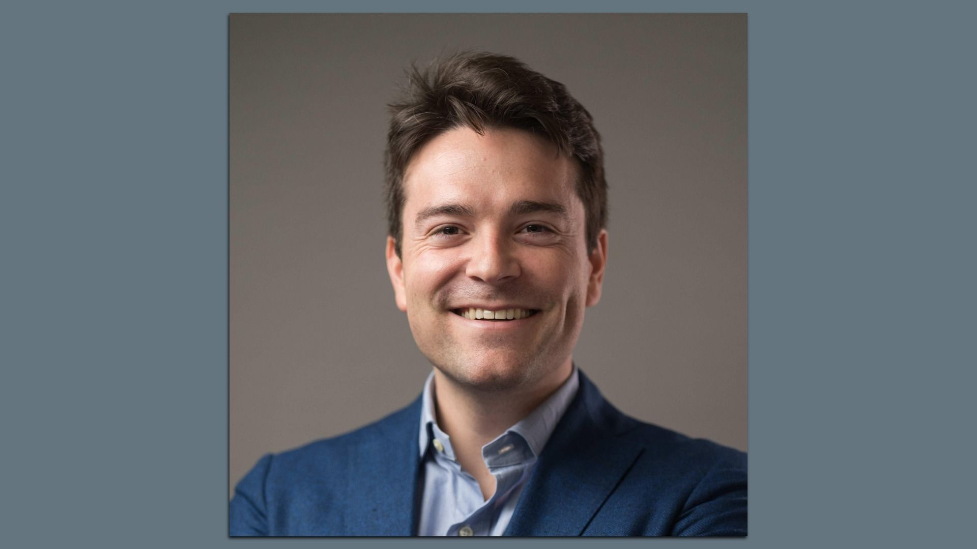 Smiling young man with dark hair wearing a blue blazer and light blue shirt, posed against a plain gray background.