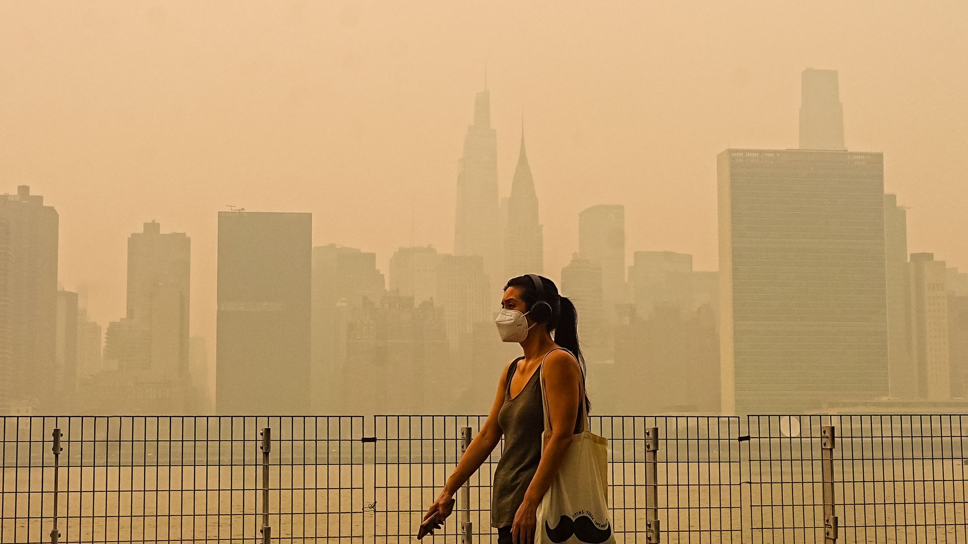 A person wearing a face mask and headphones walks along a rooftop railing, with a hazy orange city skyline and tall buildings in the background.
