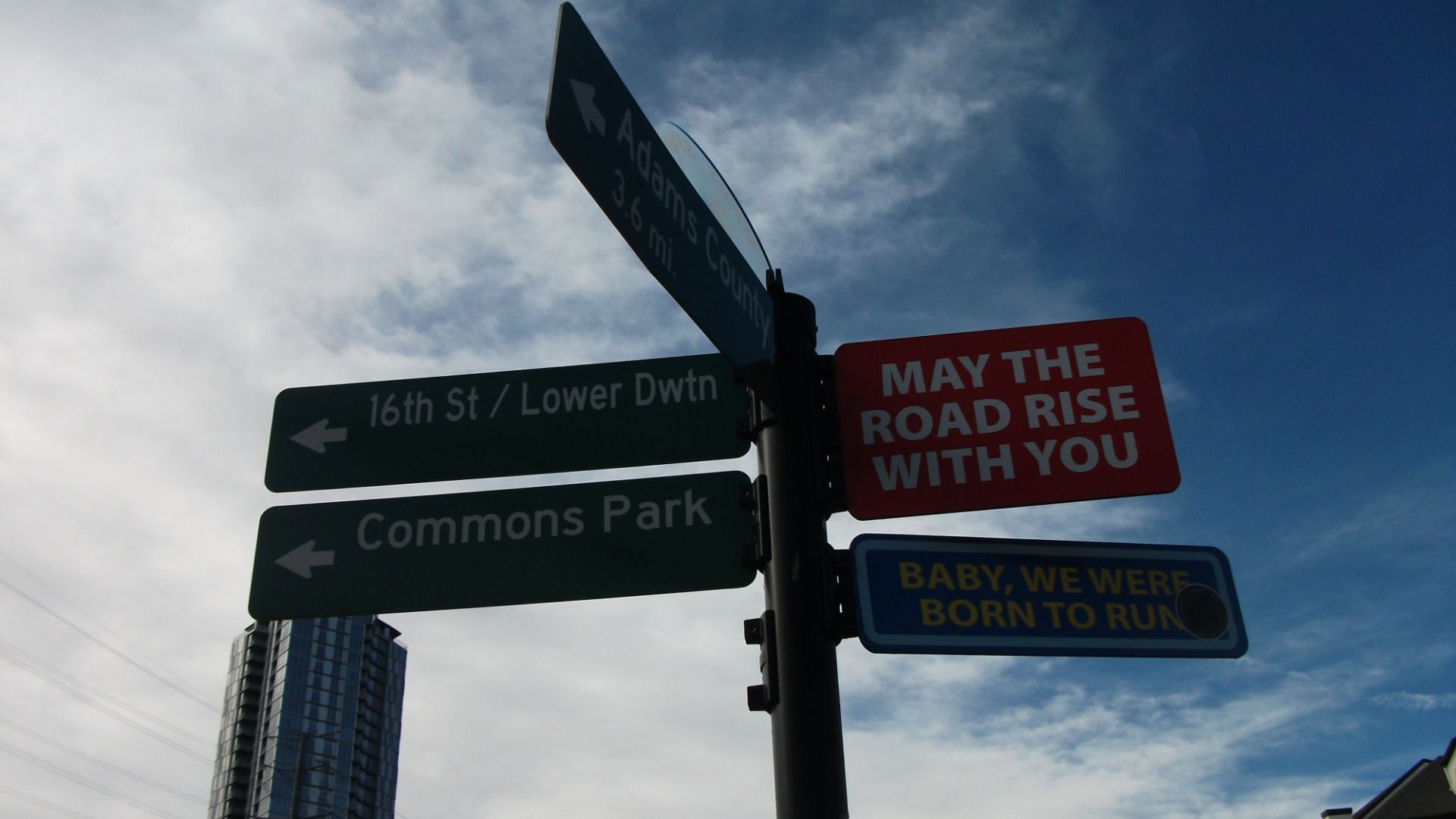 A photo of a several street signs attached to a post. One says "May the road rise to meet you." Another has an arrow and says "Commons Park."