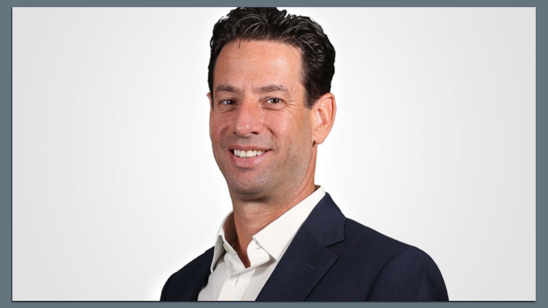 Head-and-shoulders portrait of a smiling man with dark hair, wearing a navy suit and white shirt, angled slightly to the right against a light gray gradient background.