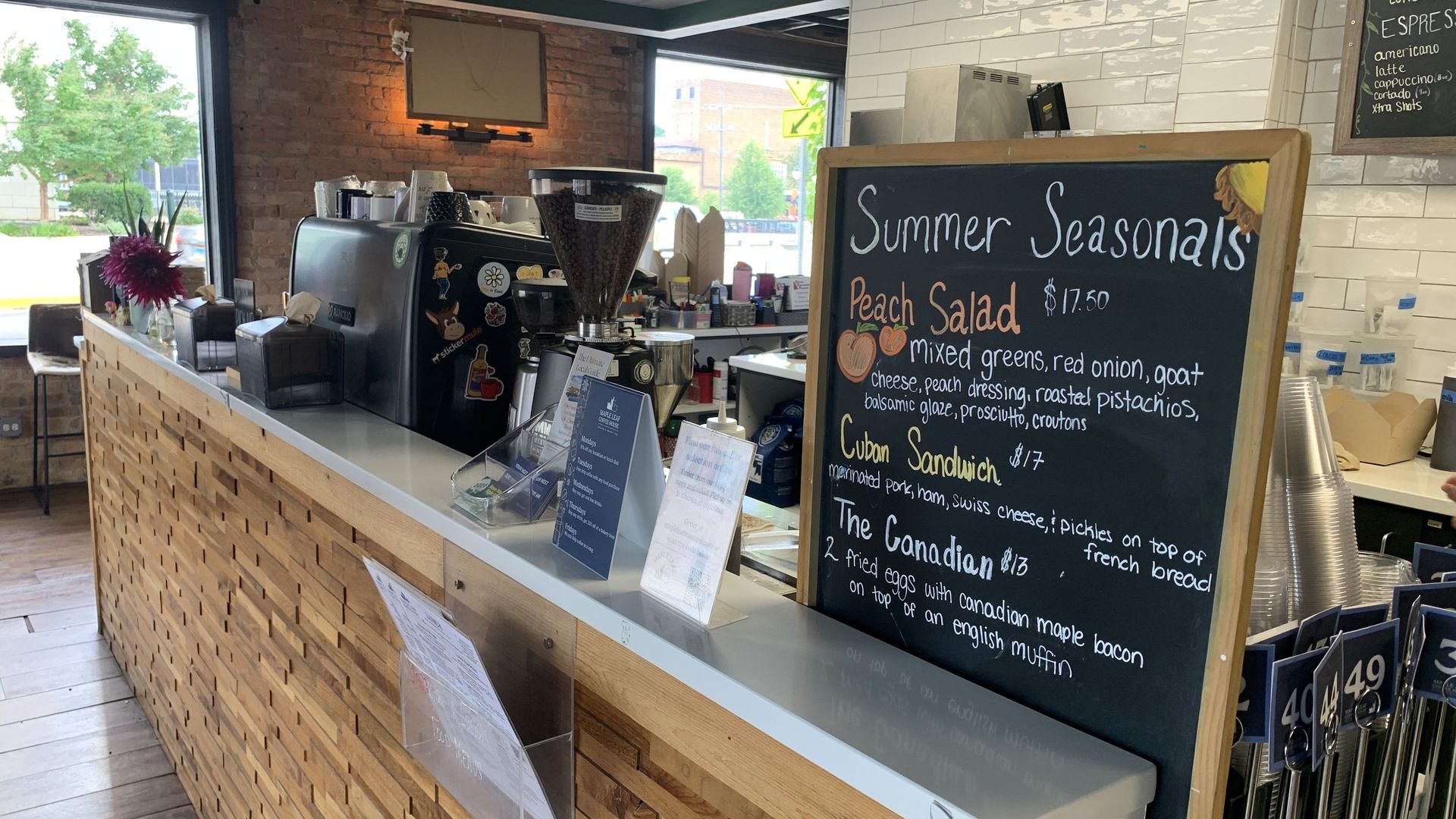 Cafe counter with a tall black chalkboard reading "Summer Seasonals" and listing Peach Salad; espresso machine, grinder, cups, and a brick wall with white subway tiles behind.