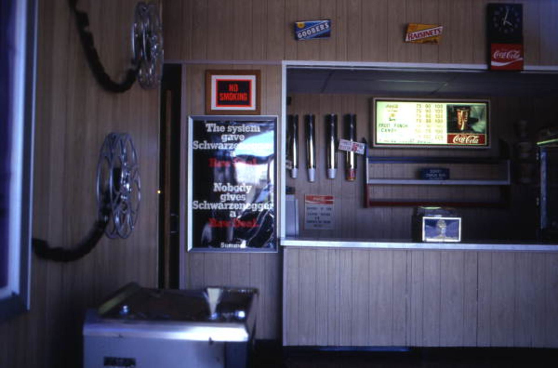 A snackbar with film reels and candy boxes hanging on the walls wood-aneled walls and a black move poster with red and white writing that reads, "The system gave Schwarzenegger a Raw Deal. Nobody gives Schwarzenegger a Raw Deal."