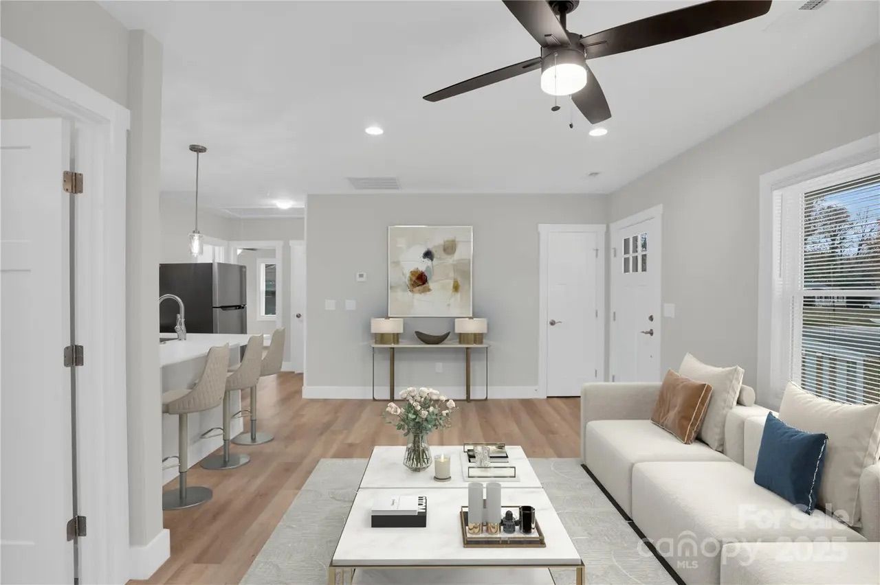 Modern living room with white sofa, blue and brown pillows, marble coffee table with flowers and candles, wood floor, gray walls, ceiling fan, and open kitchen with bar stools.