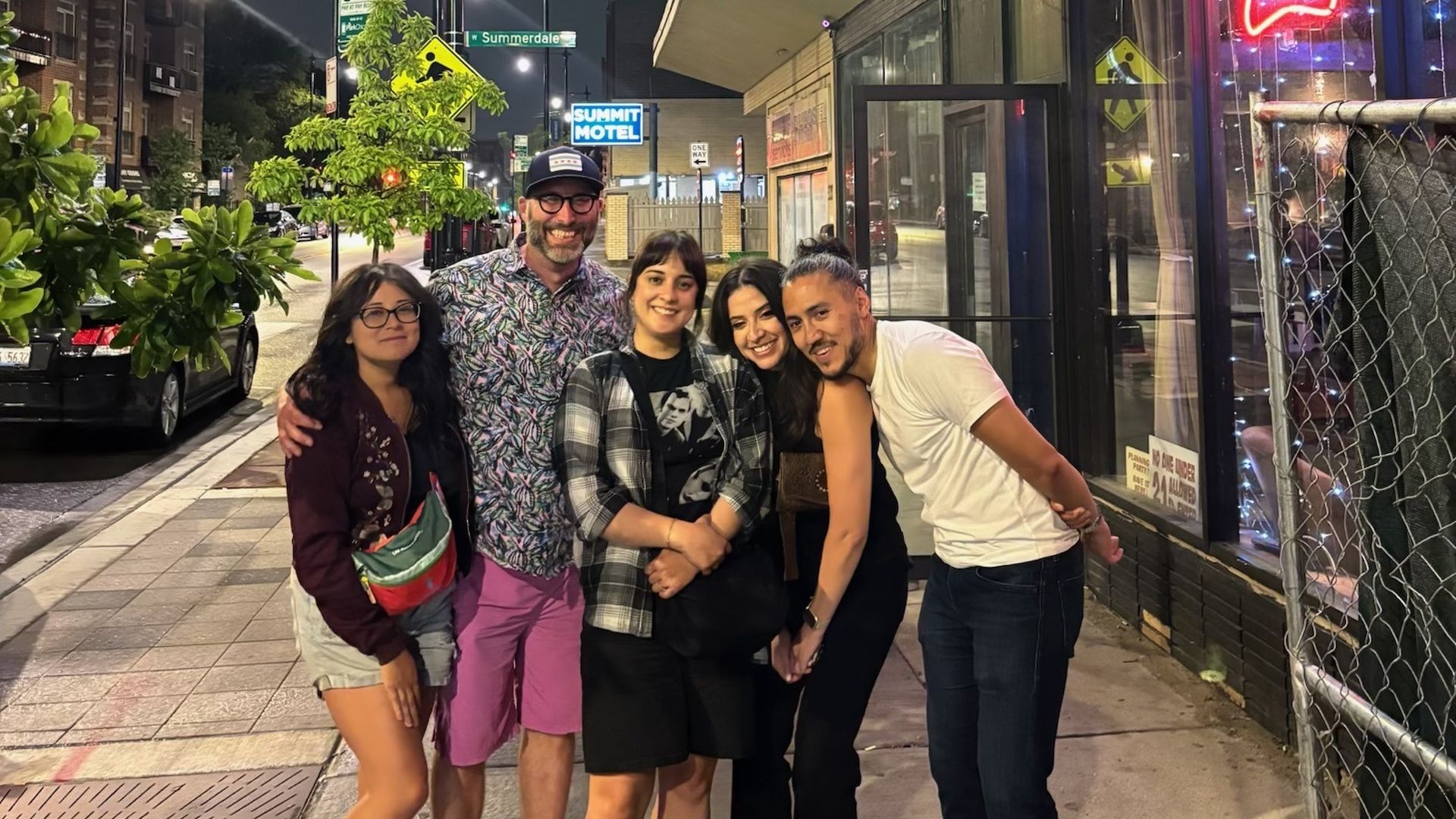 Five people smiling and posing on a city sidewalk at night near a building with neon signs and a "Summit Motel" sign visible in the background.