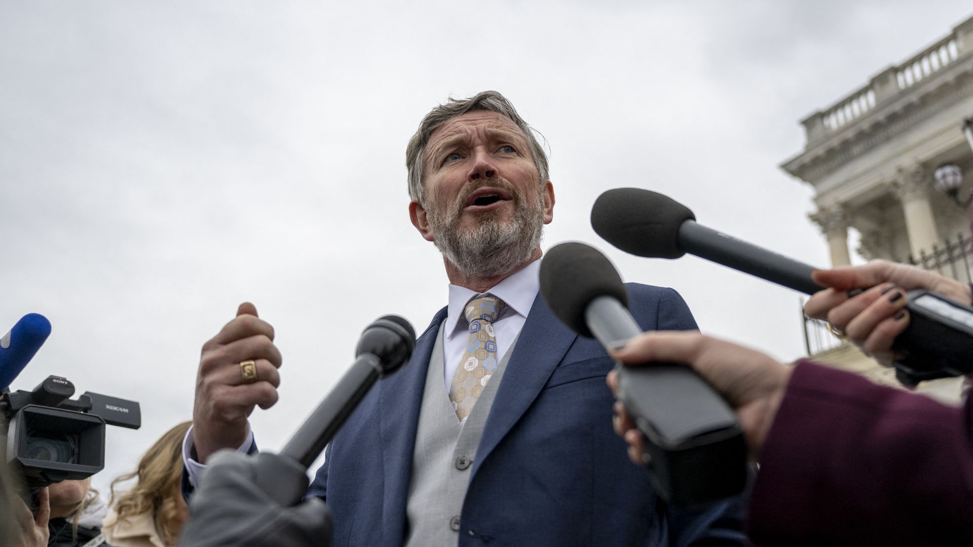 Man in a navy suit and beige patterned tie speaks to reporters; multiple handheld microphones surround him as cameras capture outside a building with columns.