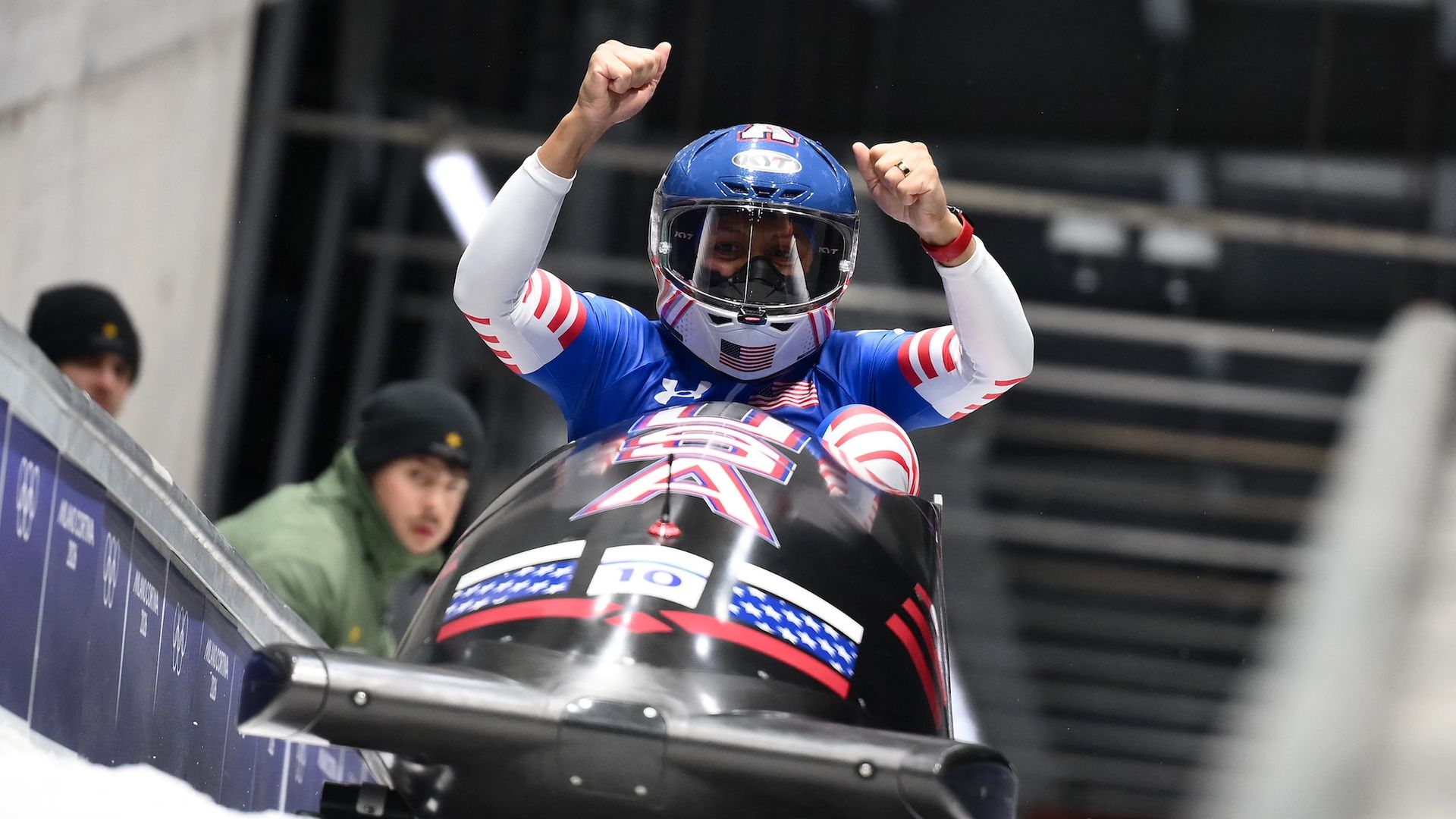A woman in a Team USA bobsled raises her arms cheering on the track after winning gold at the Olympics.