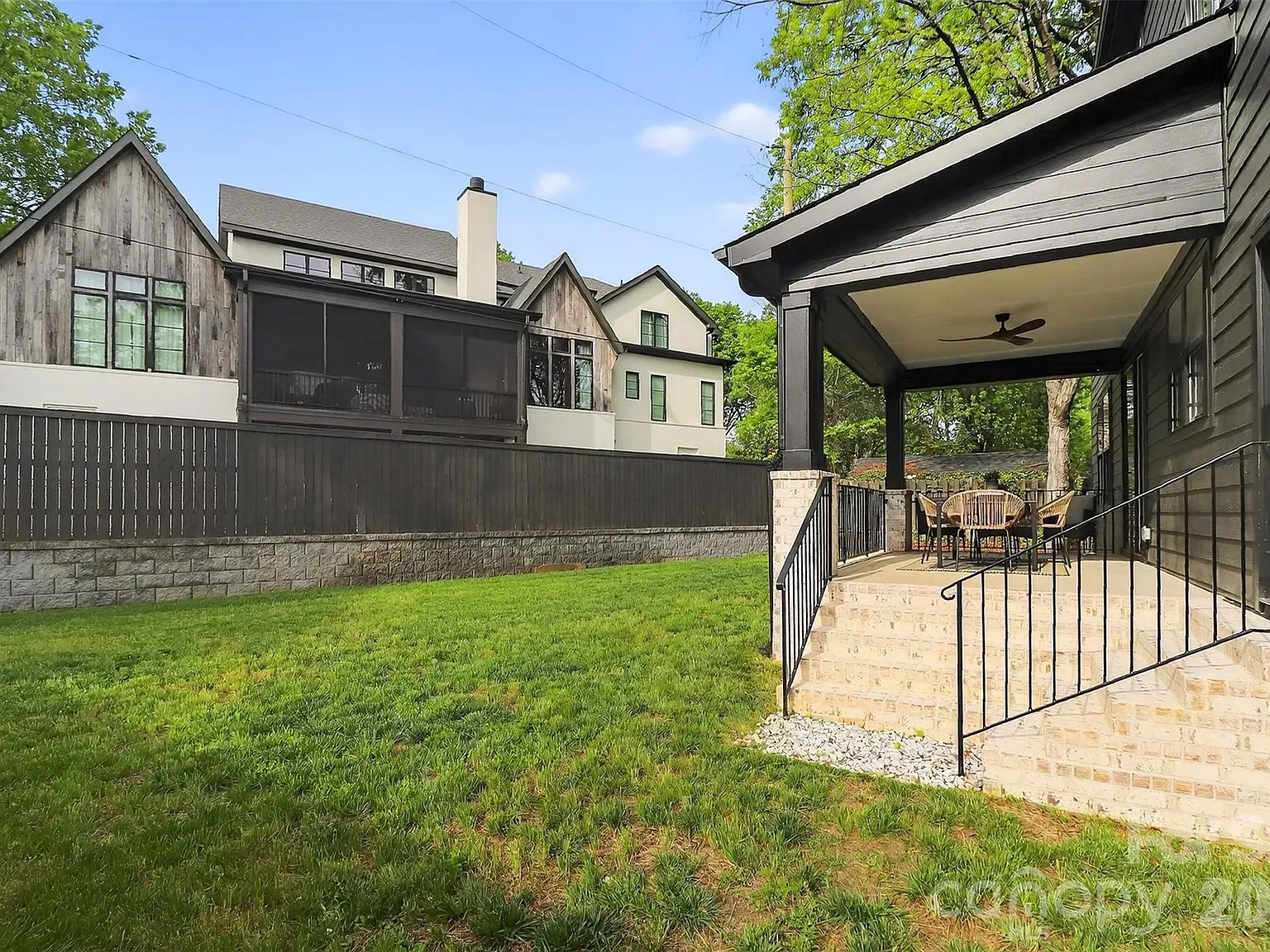 Backyard with a covered porch on the right, beige stone steps and black iron railing, a table and chairs under a ceiling fan. A green lawn and neighboring gray wooden houses in the background.