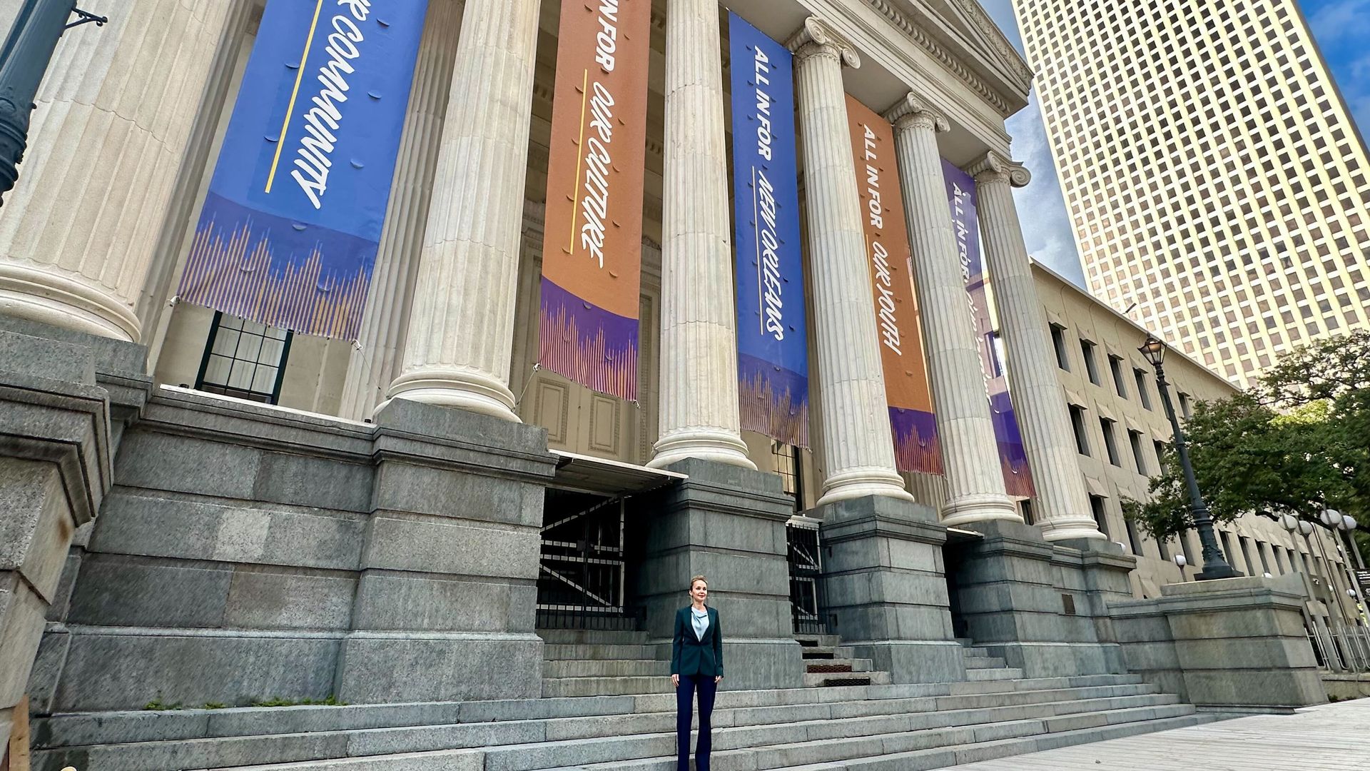 Person in formal attire standing in front of a large building with tall white columns and banners in blue, orange, and purple promoting community and culture in New Orleans.