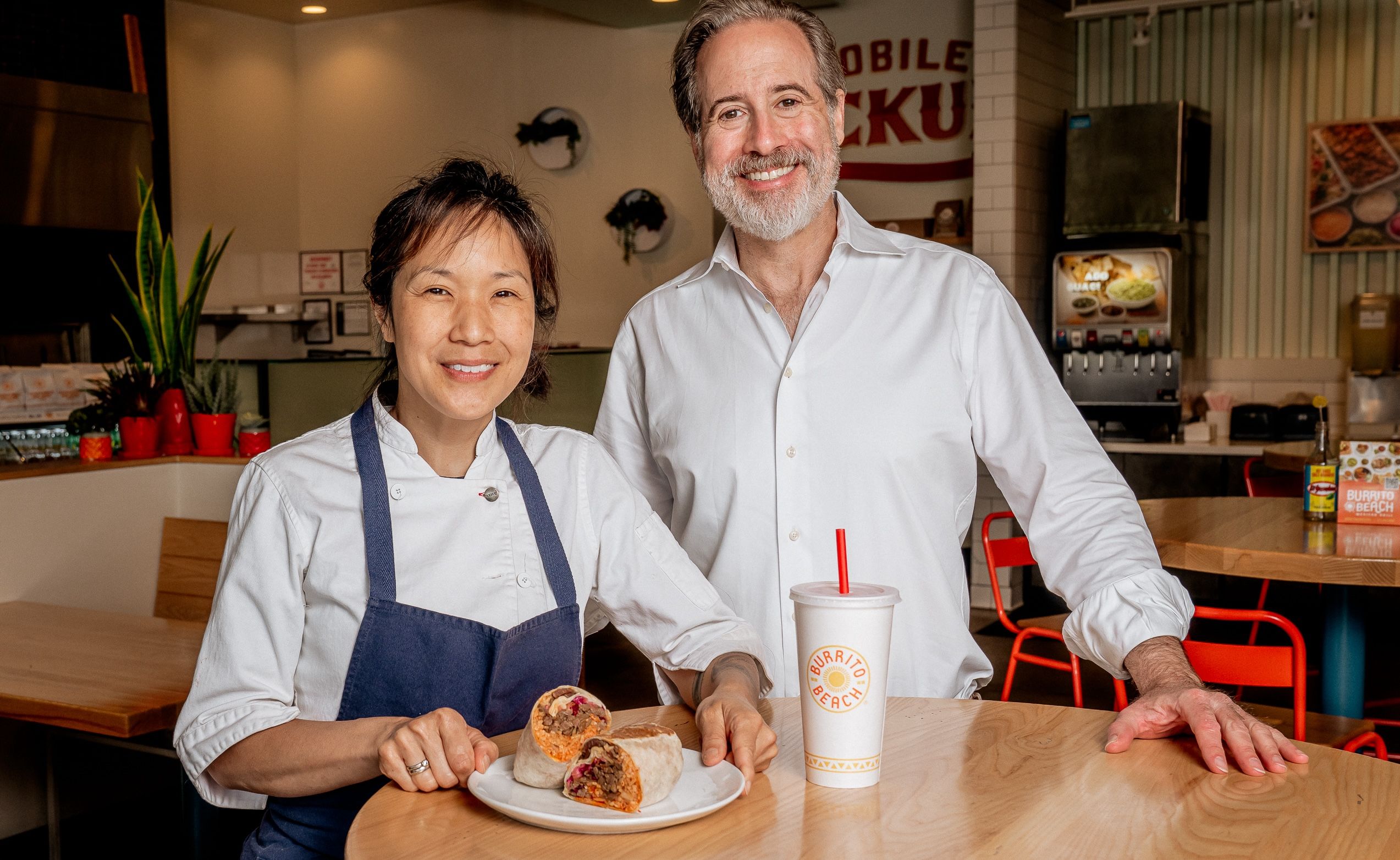 Two smiling adults, a woman in a chef coat and apron and a man in a white shirt, stand behind a wooden table in a casual restaurant with a burrito cut in half on a plate and a drink cup labeled "Burrito Beach."