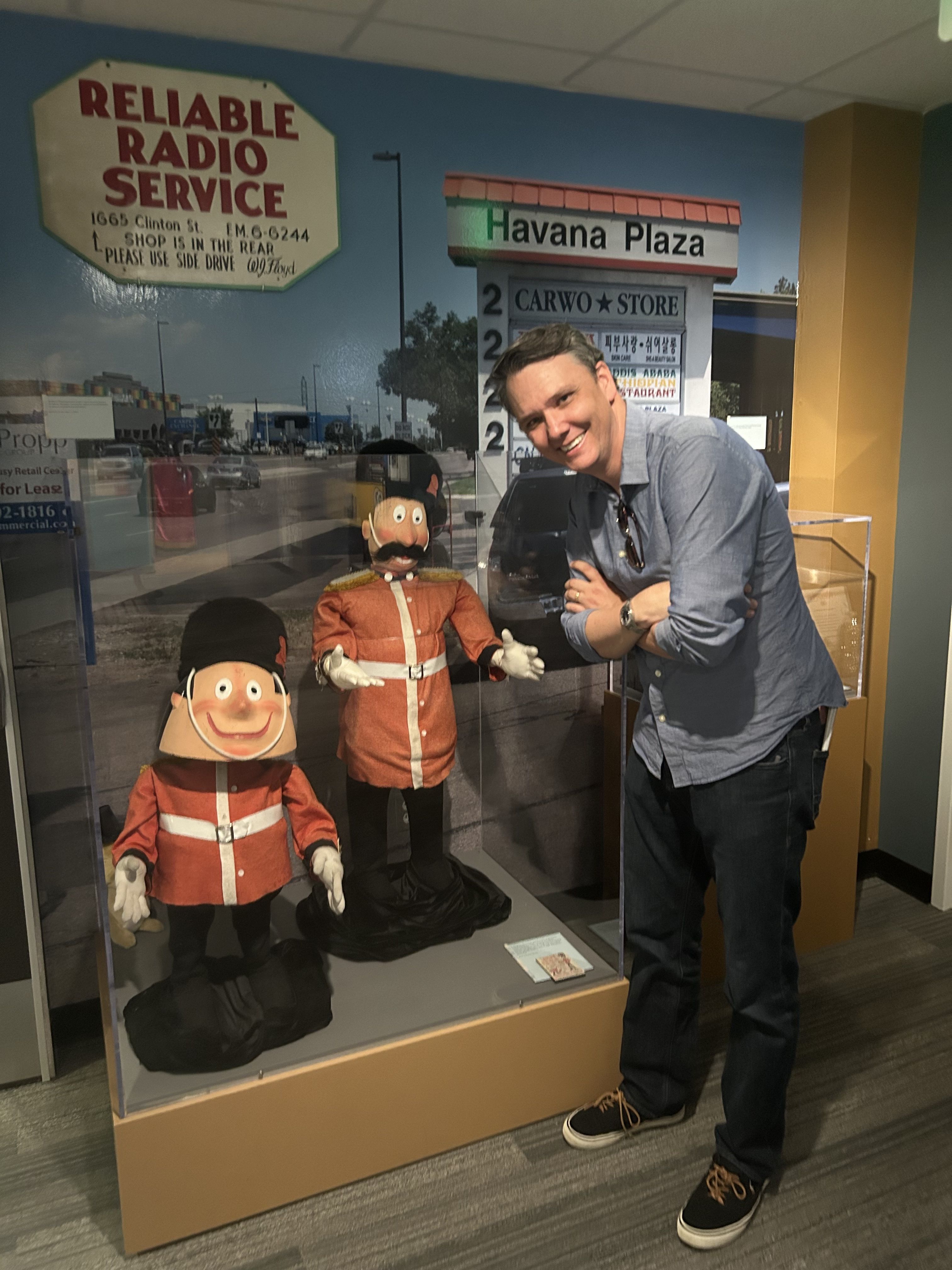 Smiling man in a blue shirt leans beside a glass display with two red-clad ventriloquist dummies; mural behind reads Reliable Radio Service and Havana Plaza.