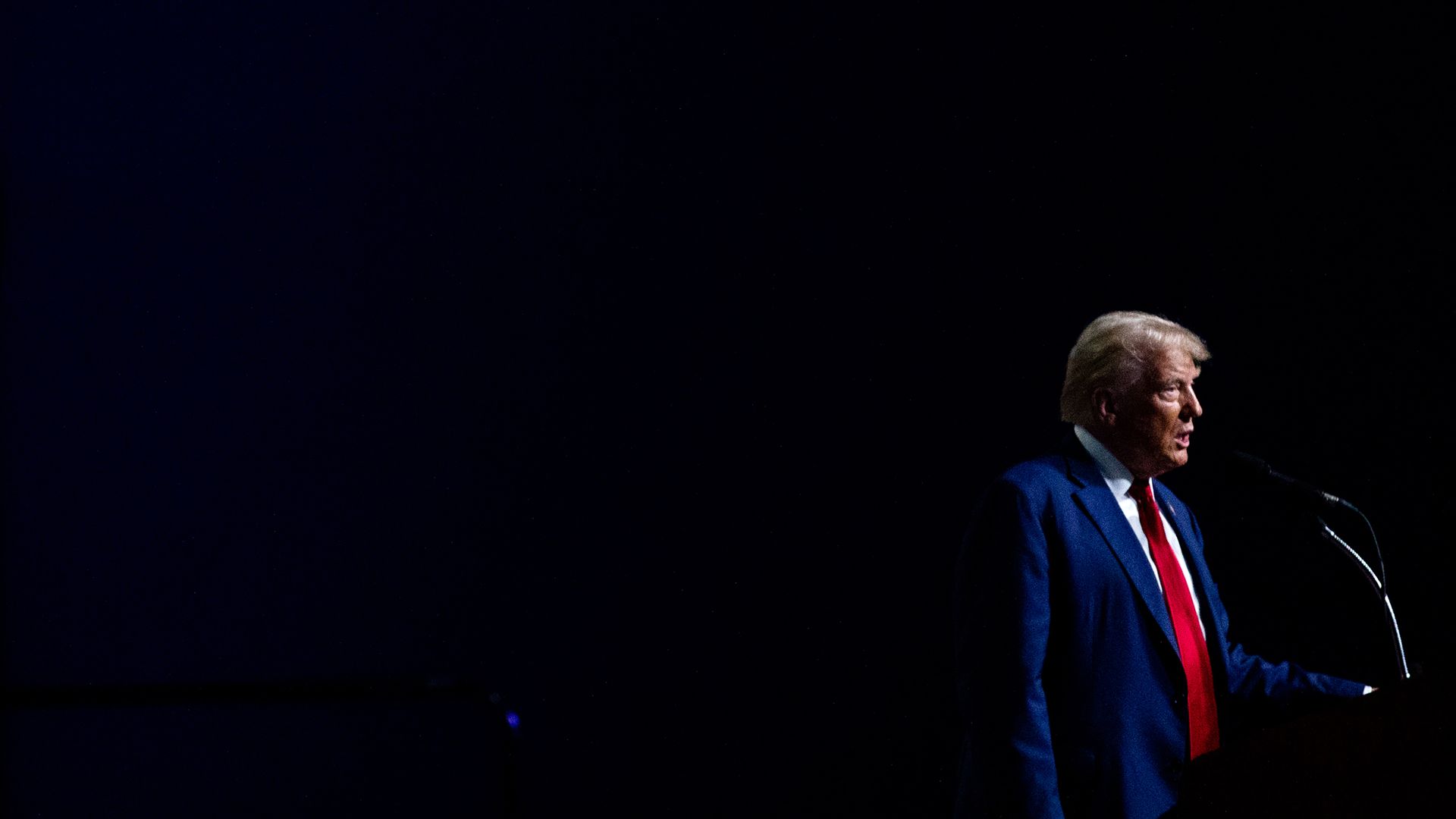 Trump speaks at a lectern. The background is black. 