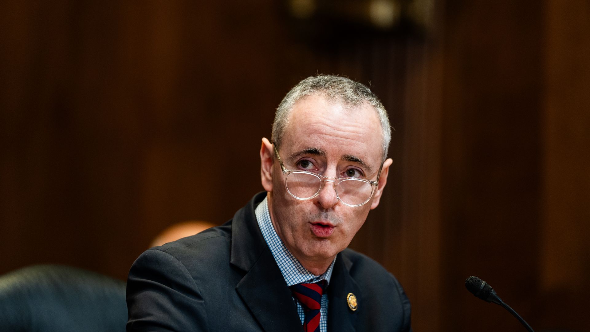 A man with short gray hair and glasses, wearing a dark suit, checkered shirt, and striped tie, speaks into a microphone against a dark brown wood-paneled background.