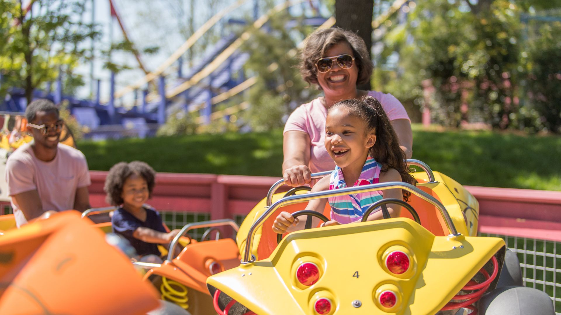 Smiling woman wearing sunglasses guides a young girl in a striped shirt riding a bright yellow amusement car; pink railing and green trees visible, with blurred riders in the background.