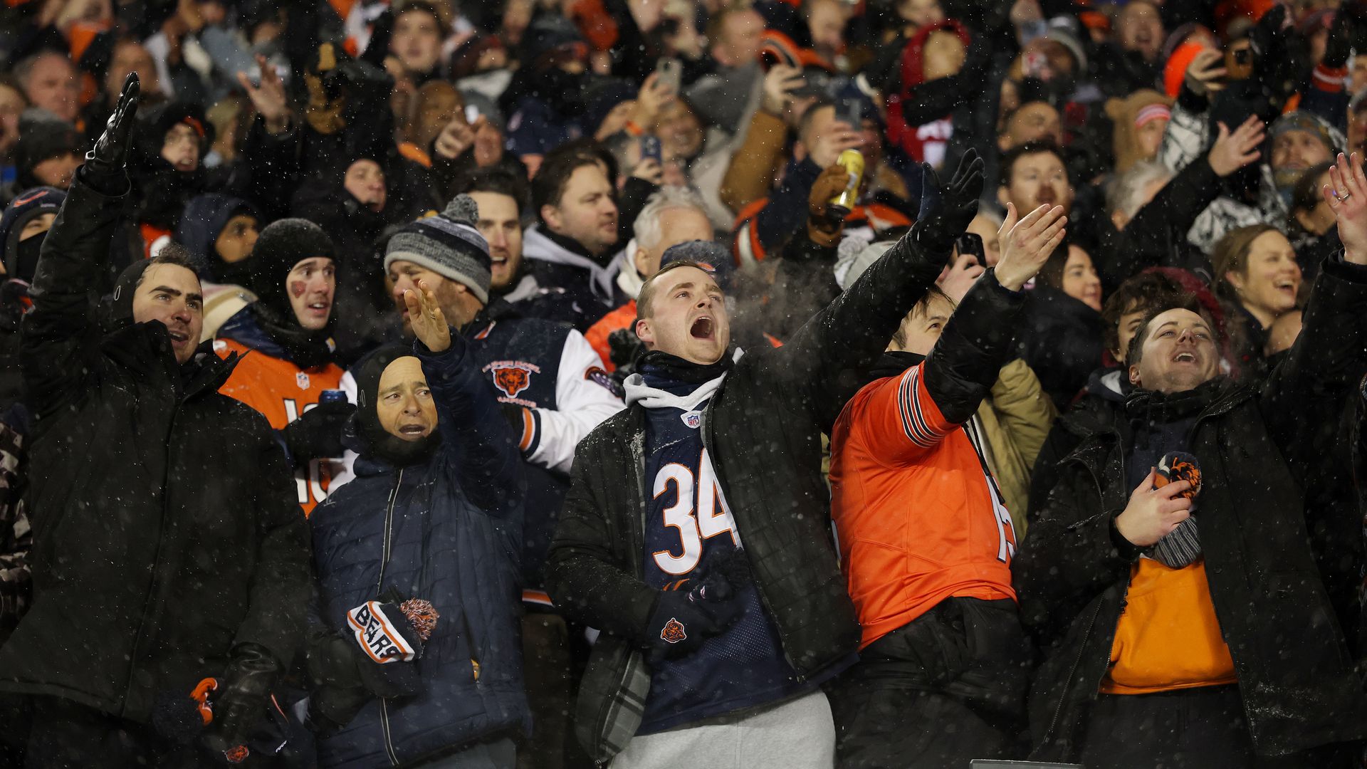 A large crowd of sports fans in Bears colors cheer with arms raised in snowfall; some hold up phones, faces excited.