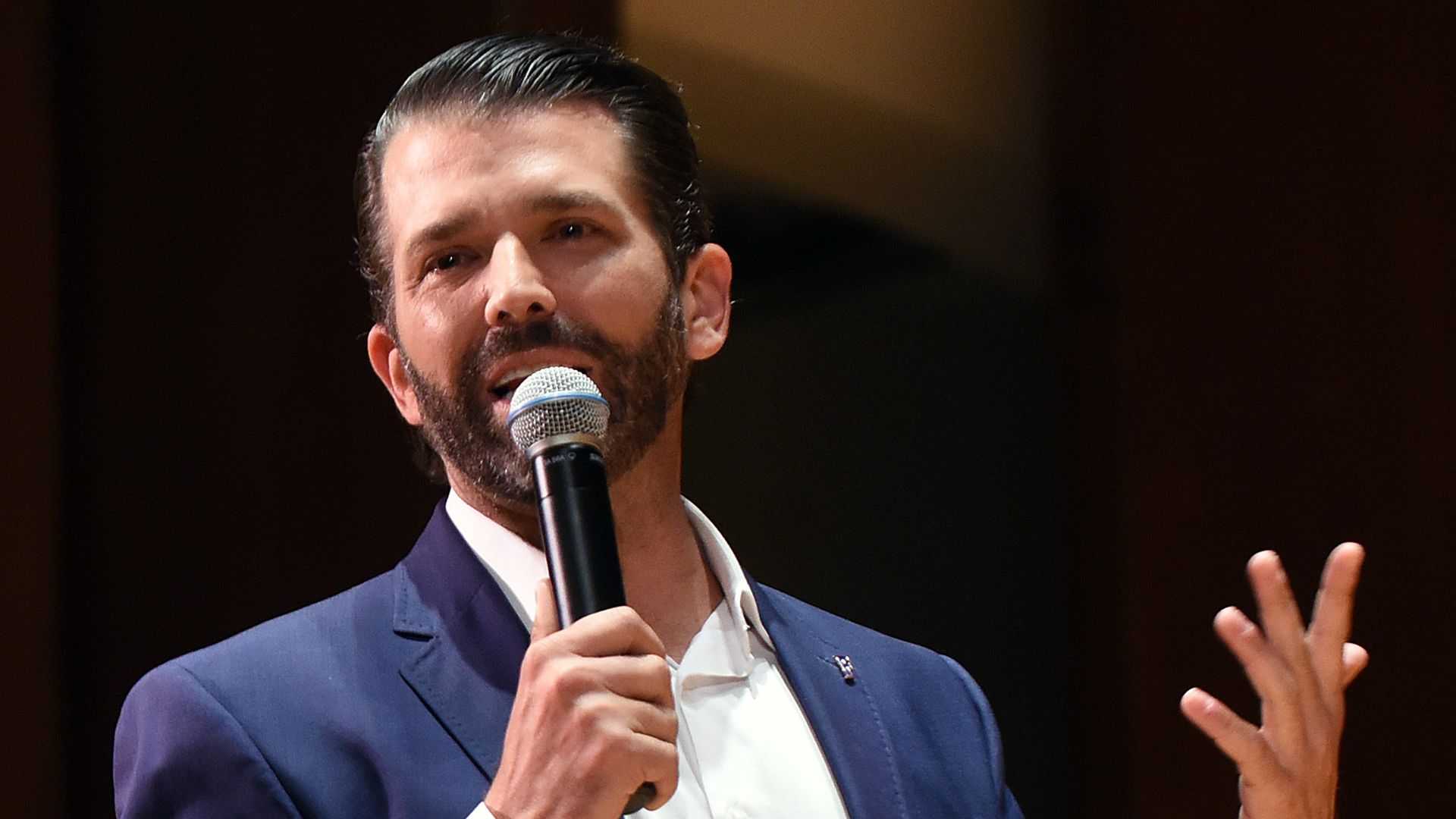 In this image, Donald Trump. Jr. stands and talks into a microphone.