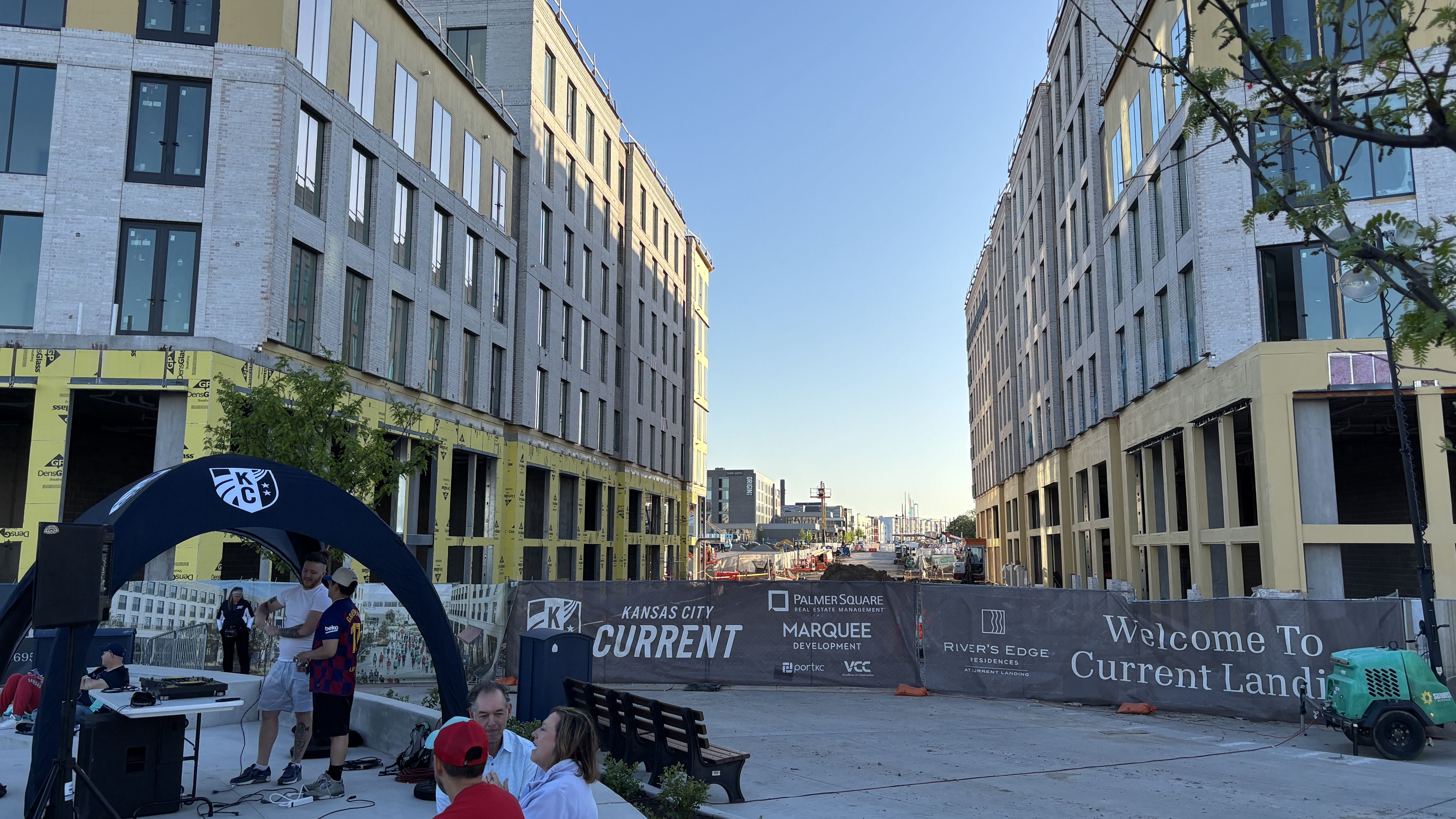 Two tall, unfinished buildings flank a wide street under a clear blue sky. A construction site with yellow sheathing and banners for Kansas City Current; a blue canopy and people gather near gear.