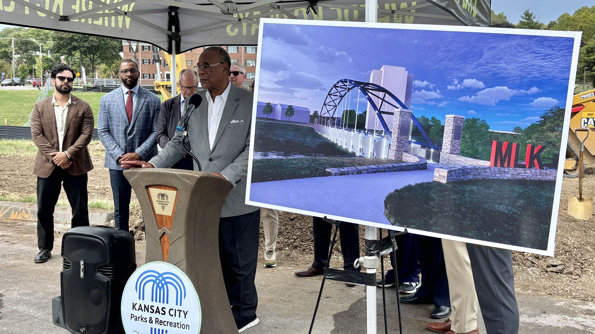 Outdoor press event under a canopy. A man speaks at a podium marked Kansas City Parks & Recreation; a large display shows a blue sky, a modern bridge, and red MLK letters.