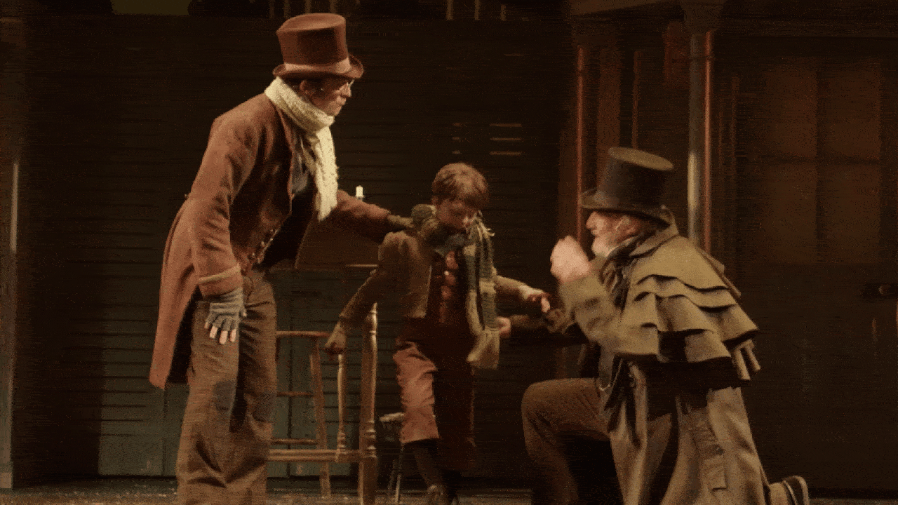 Scene with two men in brown Victorian clothing and top hats helping a young boy in a brown outfit and scarf step down from a chair on a dimly lit stage.