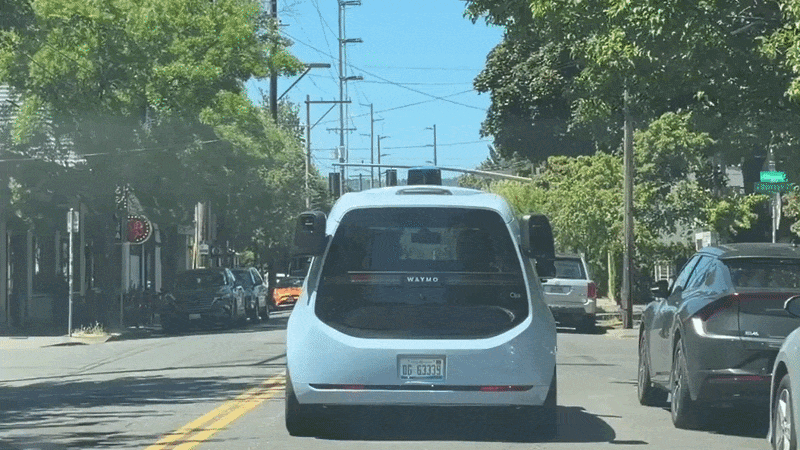A blue van driving down a city street with cars parked alongside. The van has cameras on its rear sides and on top of the car.