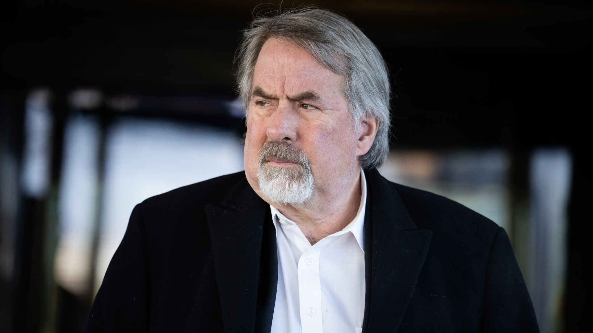 Man with gray hair and beard, wearing a white shirt and black coat, looking to his left with a serious expression, blurred dark background.