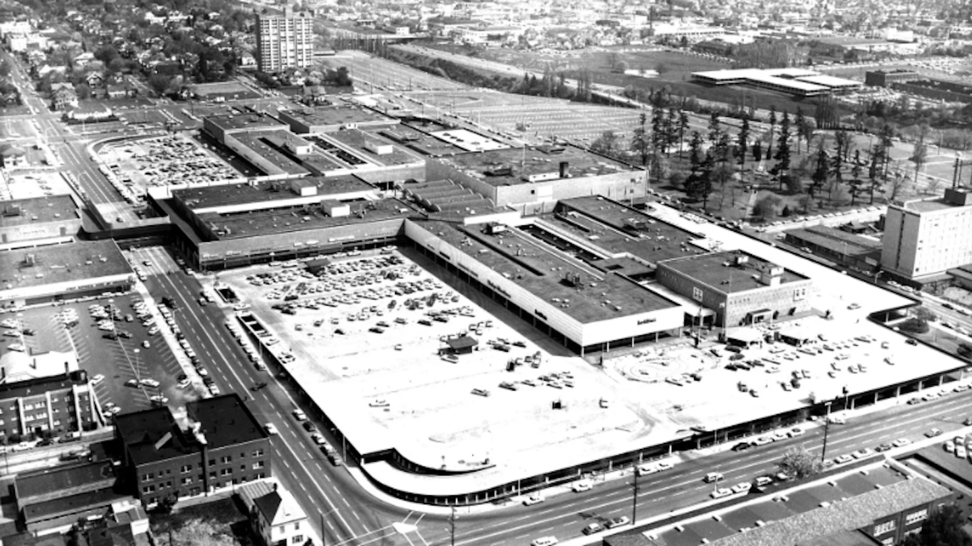 Black-and-white aerial view of a large shopping mall complex with a curved corner, extensive parking lots full of cars, surrounding streets, and nearby low-rise buildings.