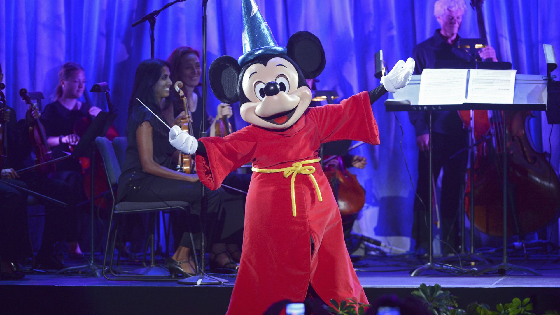 Mickey Mouse in red sorcerer's robe and blue hat conducts orchestra with musicians holding string instruments on a stage lit with blue light.
