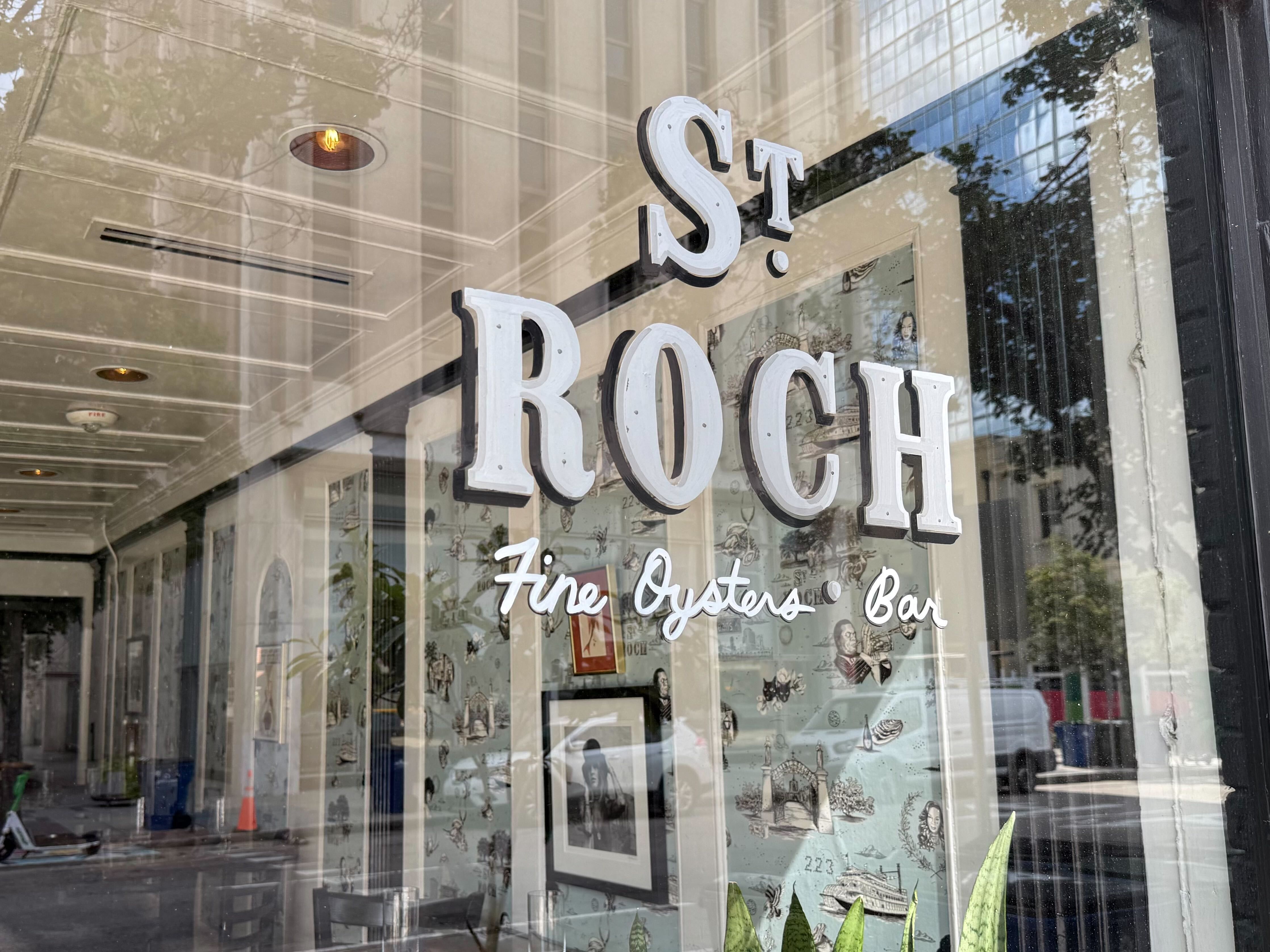 Window of St. Roch Fine Oysters Bar showing white and black lettering, vintage wallpaper with sketches inside, and reflections of street and trees outside.