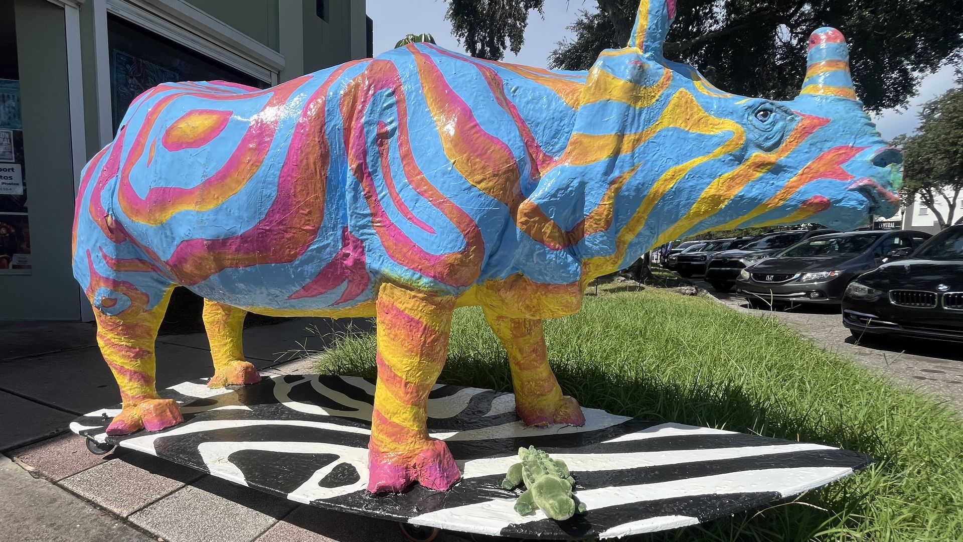 Colorful blue rhinoceros statue with pink, orange, and yellow swirls stands on a black-and-white zebra-striped pedestal outside a storefront, with trees and parked cars in the background.