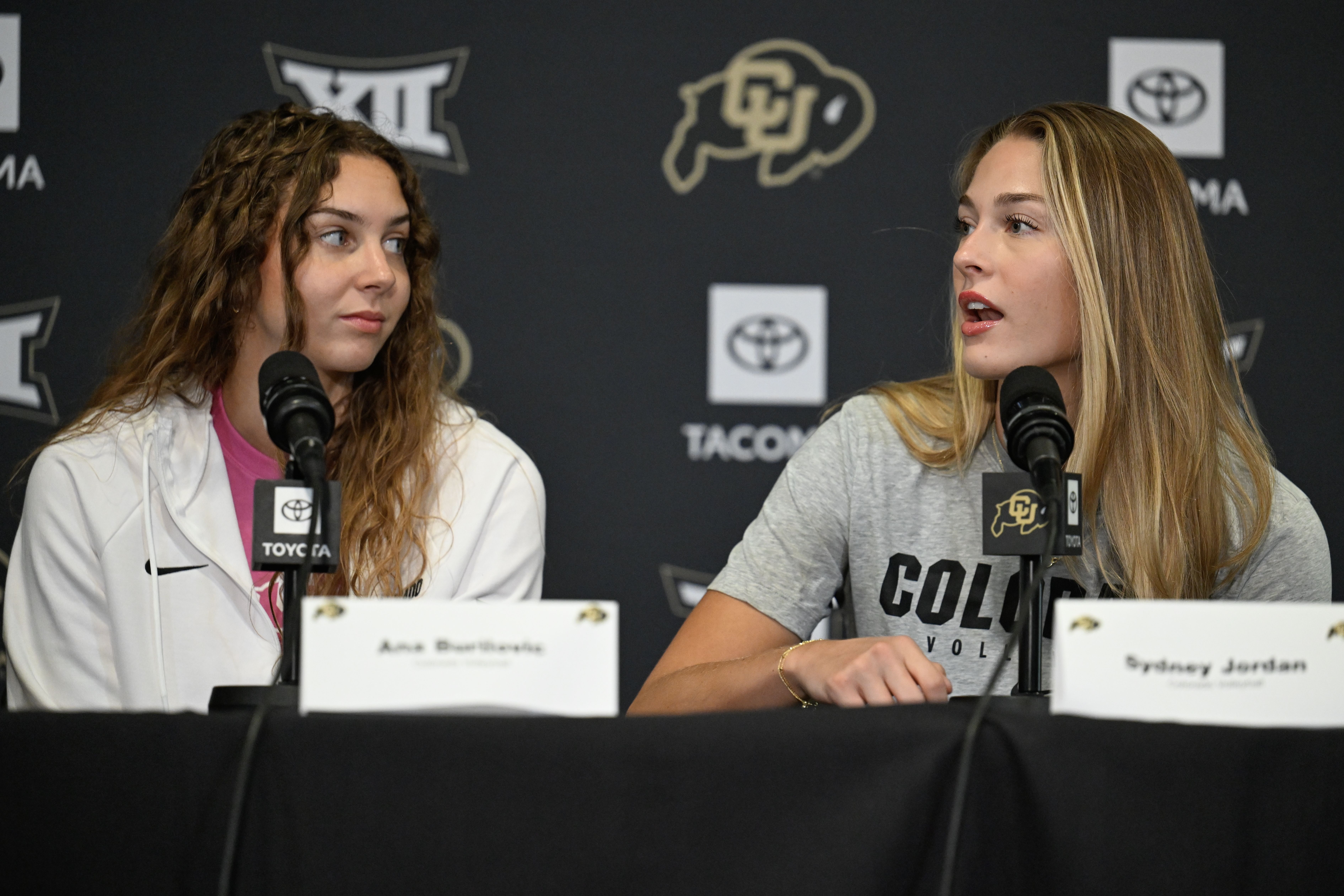 Two female athletes at a press conference, seated with microphones. Left: curly brown hair in a white jacket; right: long blond hair in a gray shirt. Backdrop features logos and a buffalo emblem.