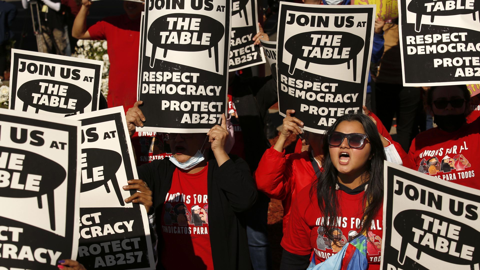 Protesters dressed in red hold large signs that say "join us at the table, respect democracy"