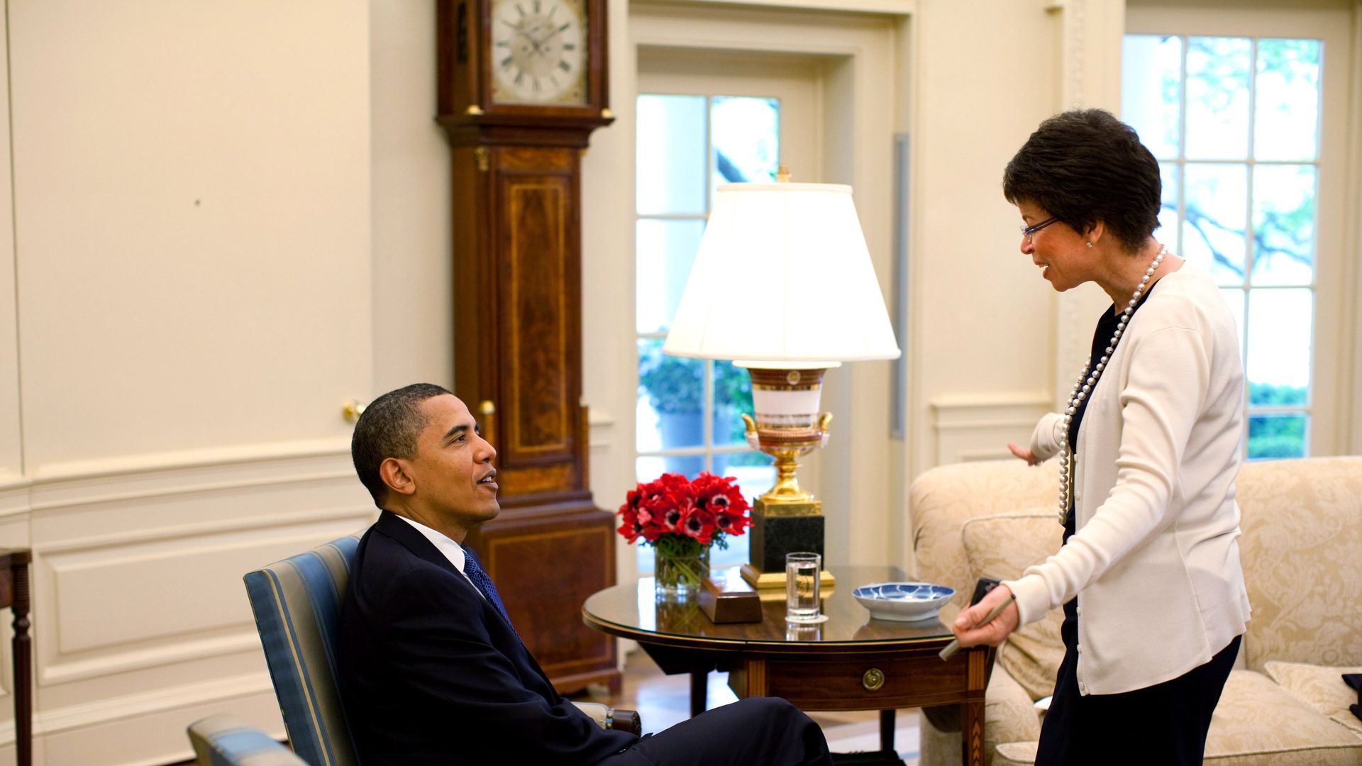 In this image, Obama is sitting on an armchair in the Oval Office while Valerie stands in a white cardigan and speaks to him.