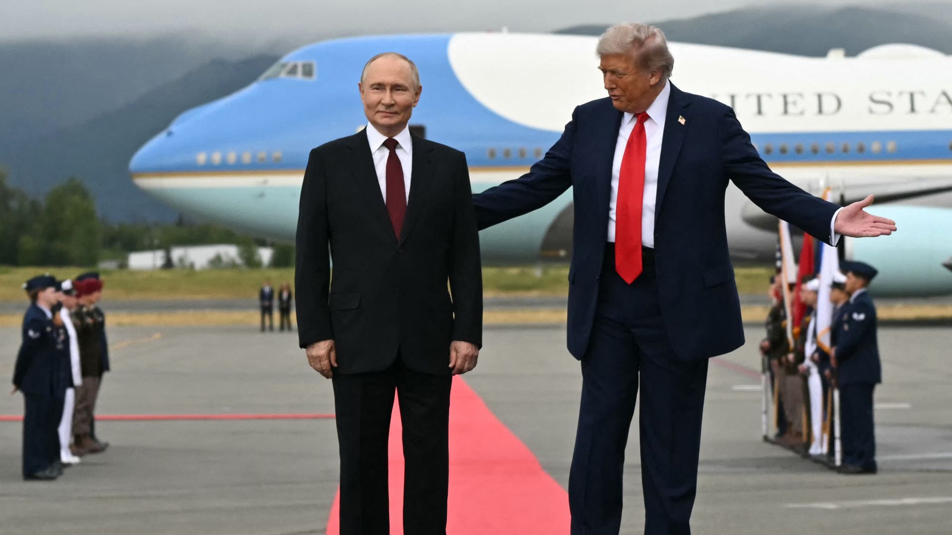 Two men in suits stand on a red carpet in front of a blue and white airplane. The man on the right gestures with open arms toward the man on the left. Military personnel stand in formation nearby.