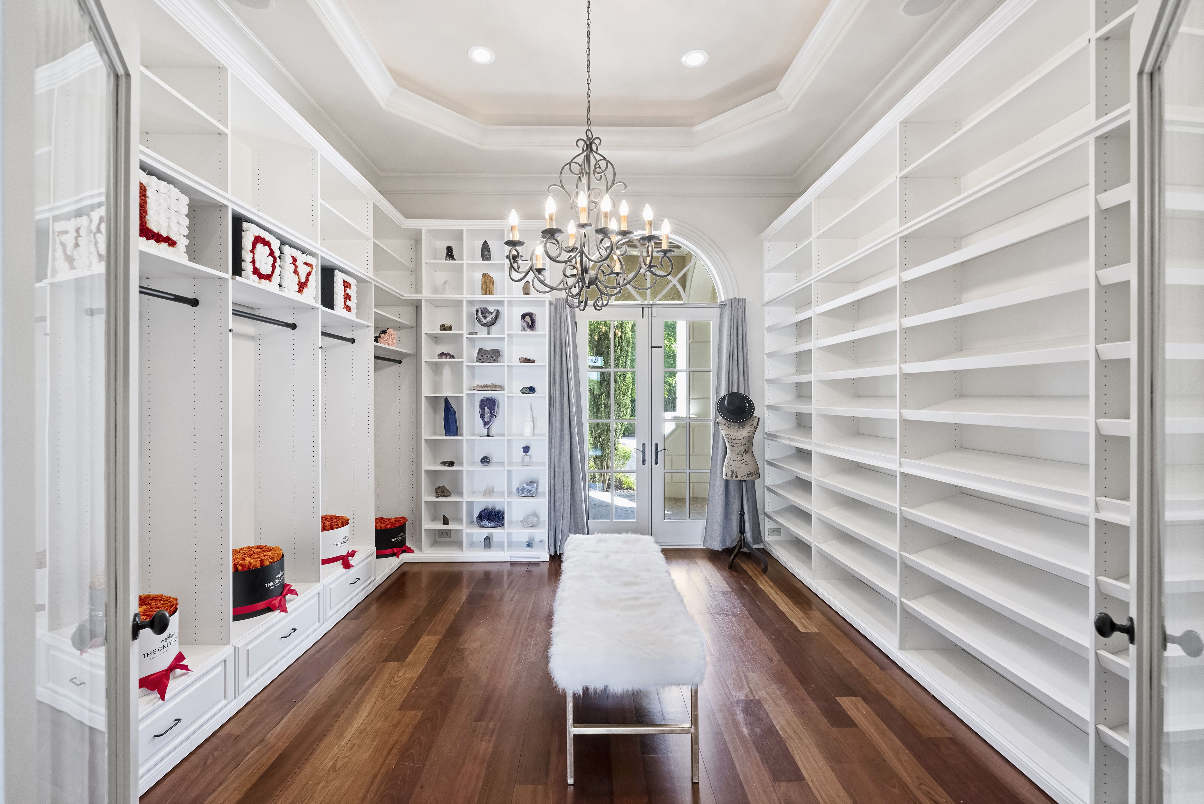 Bright white walk-in closet with tall empty shelving along both walls, a metal chandelier overhead, a white fur-covered bench in the center, and glass French doors at the far end revealing greenery; left shelves spell LOVE.