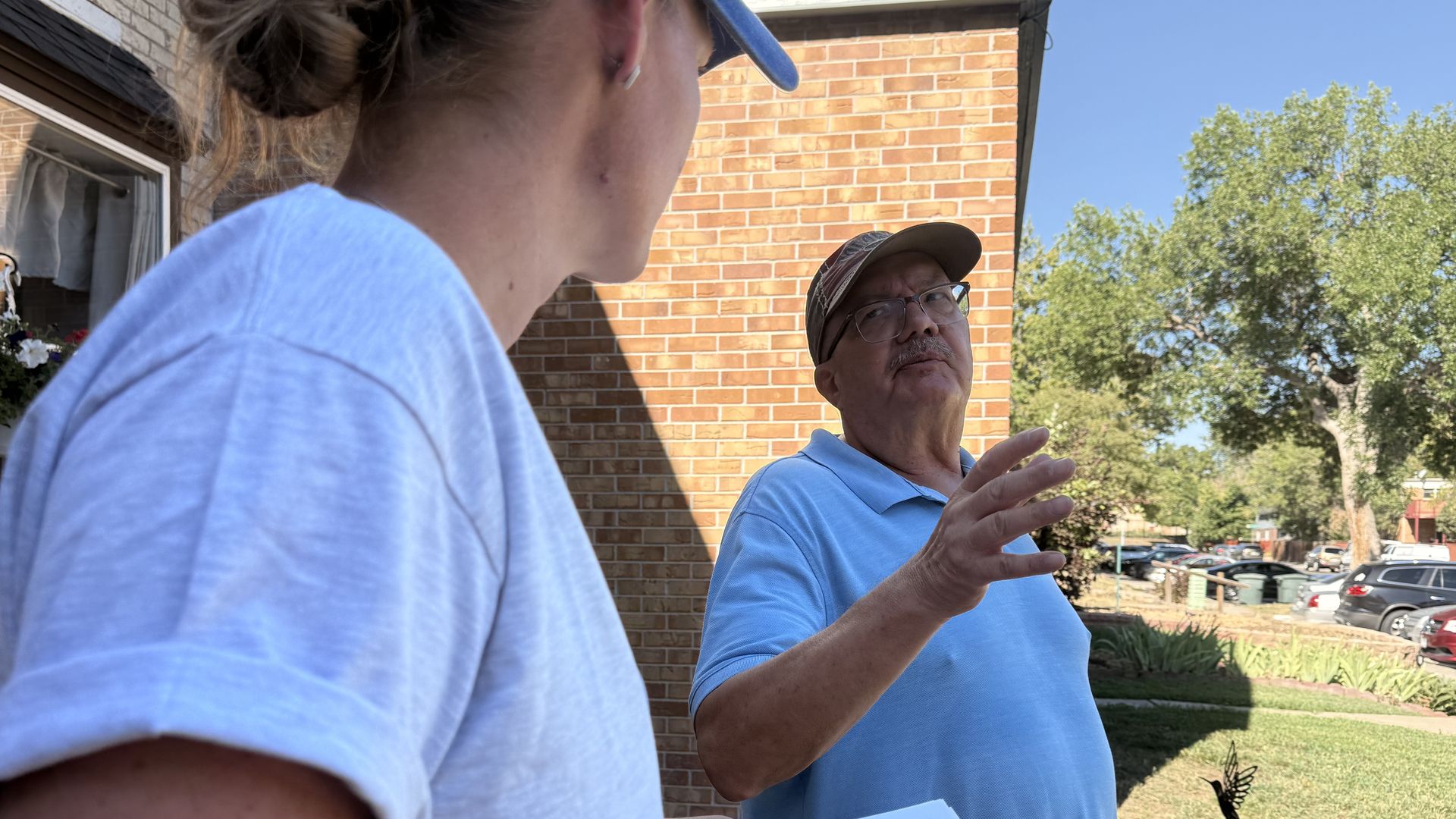 Two people talk outside a brick building. A man in a blue polo and cap gestures; another person in a light blue shirt holds papers. Trees and cars in the background.