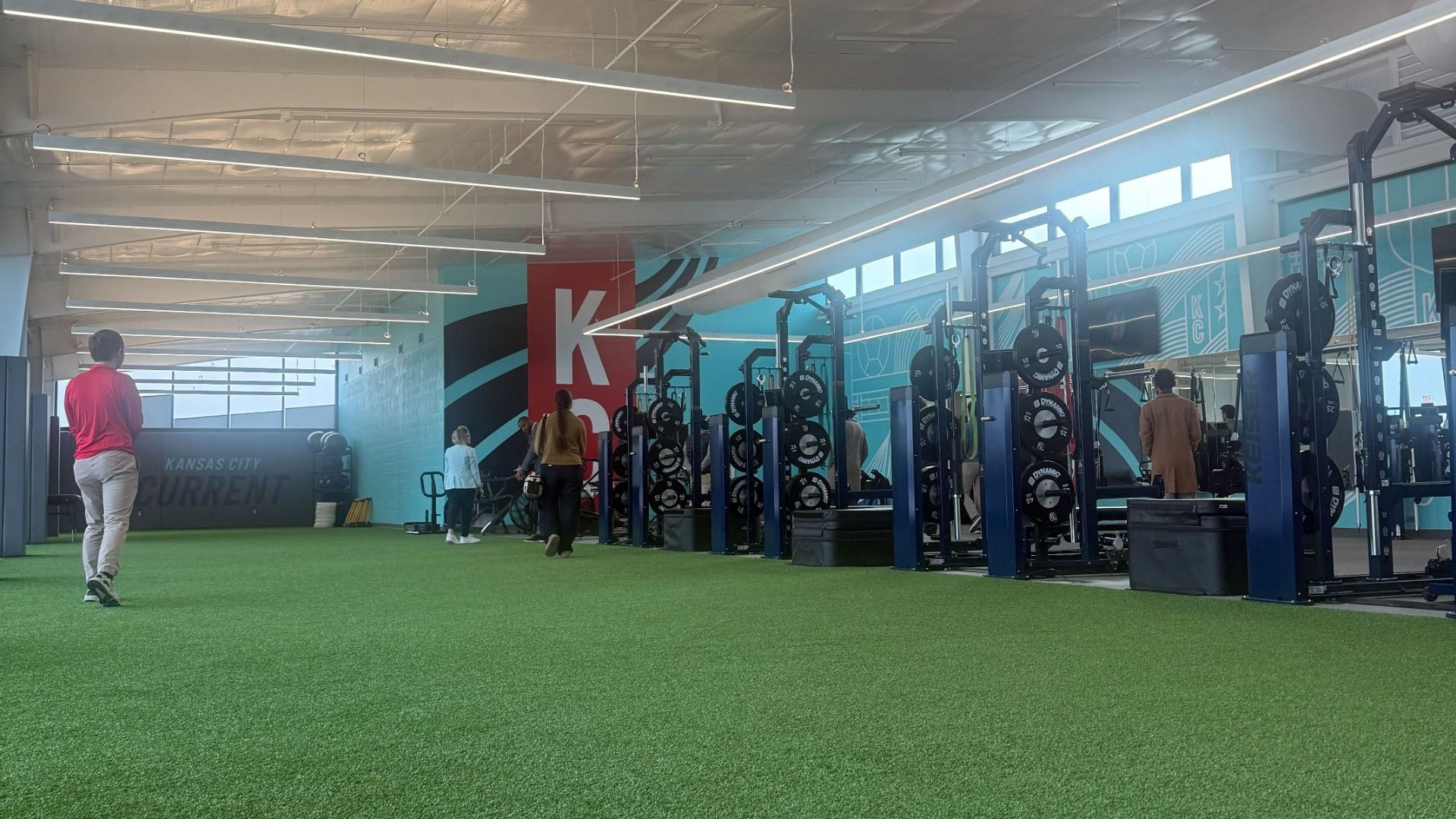 Interior of a gym with green turf floor, weight racks with black and white plates, and people walking around. Walls painted blue with Kansas City Current logo and large red "KC".