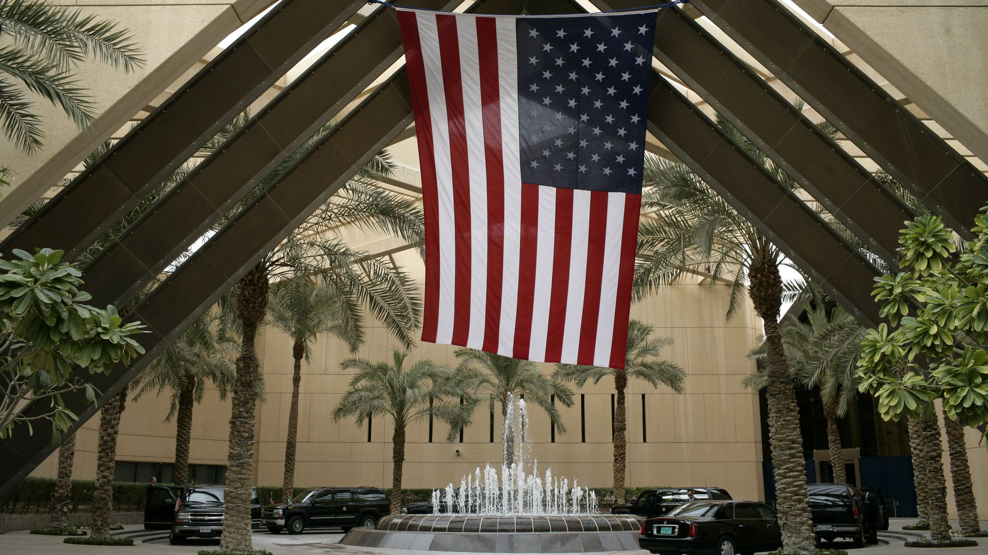 Large American flag hanging upside down inside a courtyard with palm trees, a water fountain, beige buildings, and several black vehicles parked around.