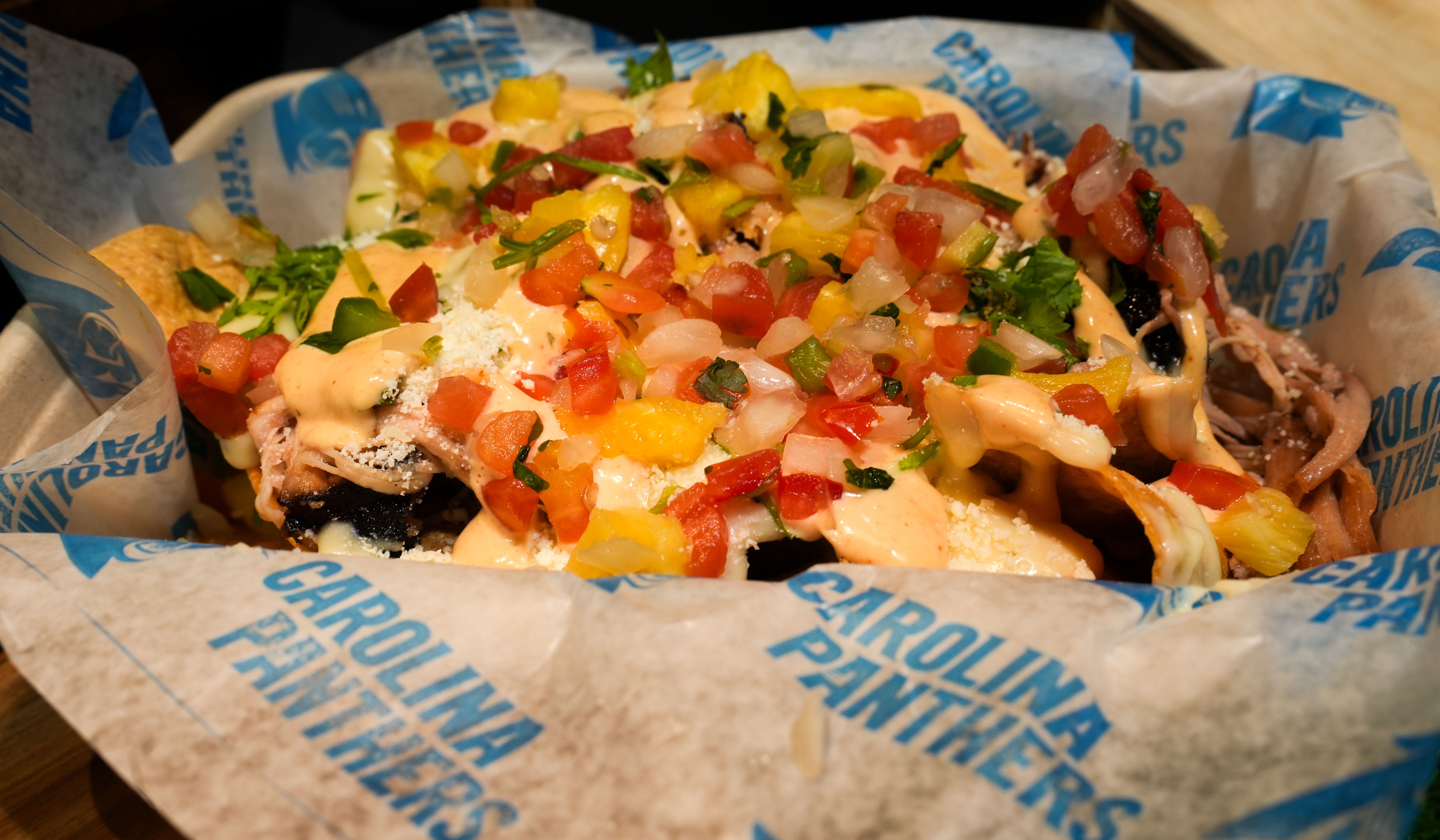 Basket of loaded nachos with diced tomatoes, onions, yellow peppers, cilantro, cheese, and creamy orange sauce on paper branded with Carolina Panthers.