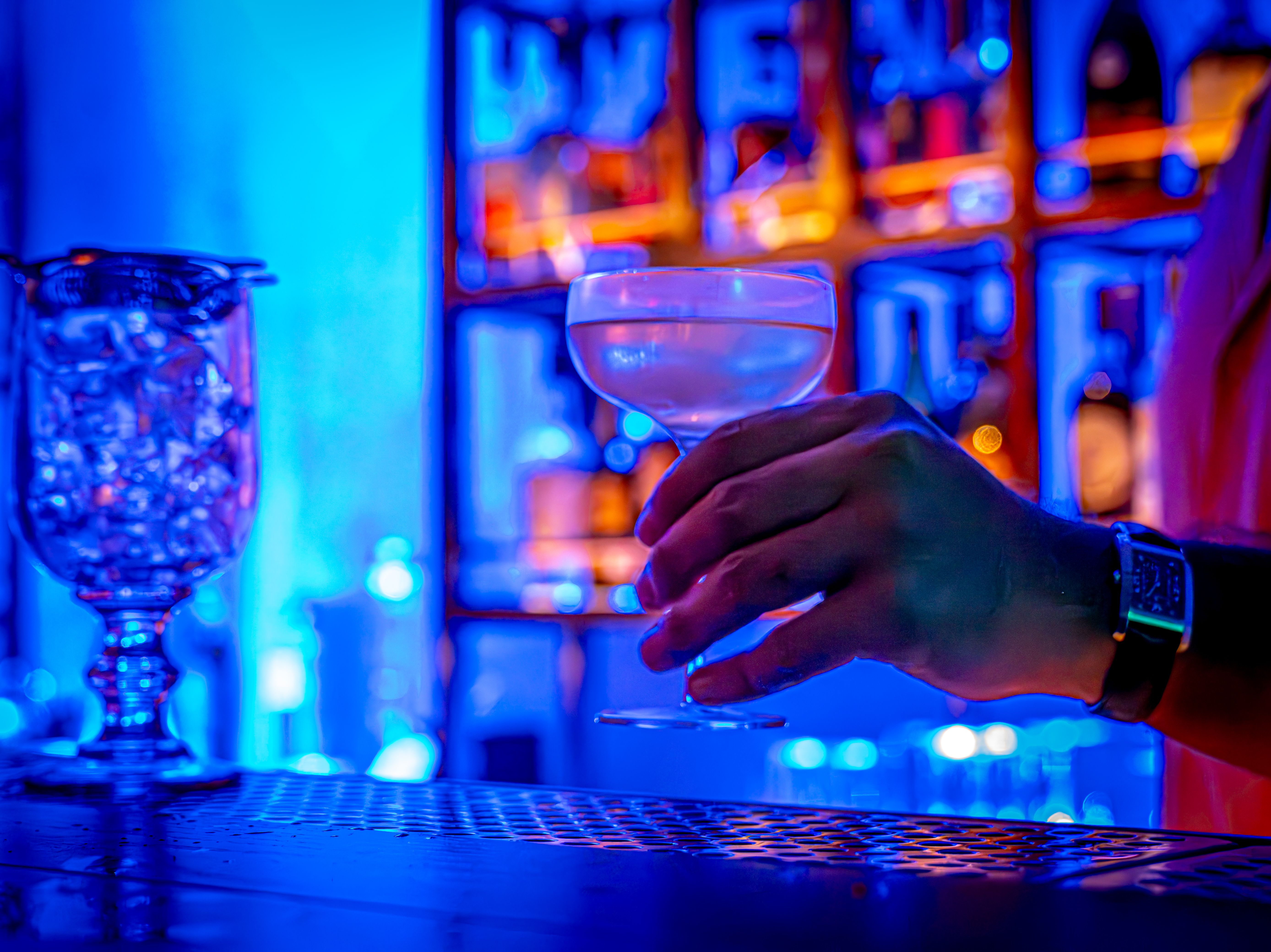 Hand holding a clear cocktail glass with a drink against a blue-lit bar background, with a large jar of ice cubes on the left and blurred shelves of bottles behind.