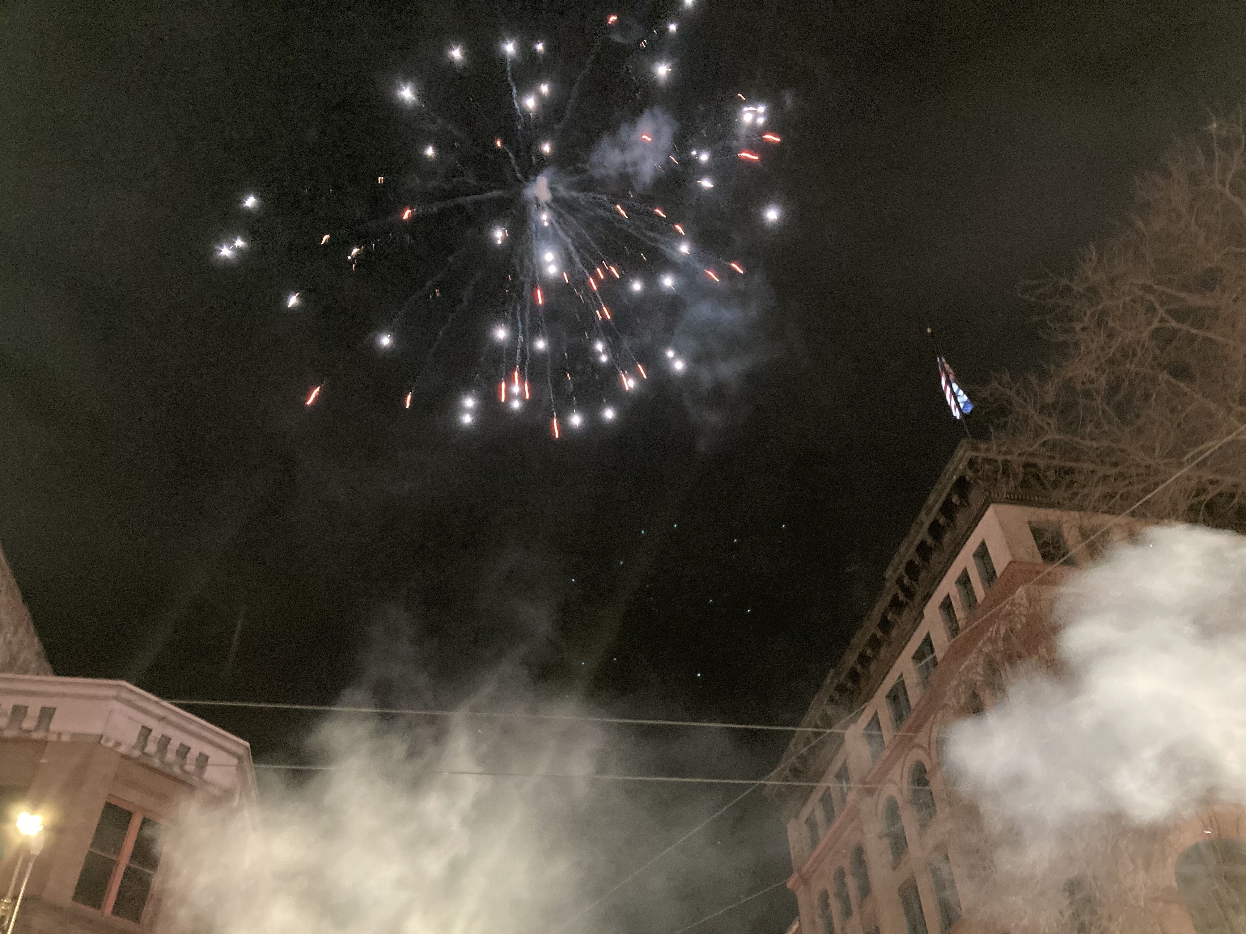 Fireworks with white and red sparks burst in the night sky above buildings, with smoke and an American flag visible on one building's roof.