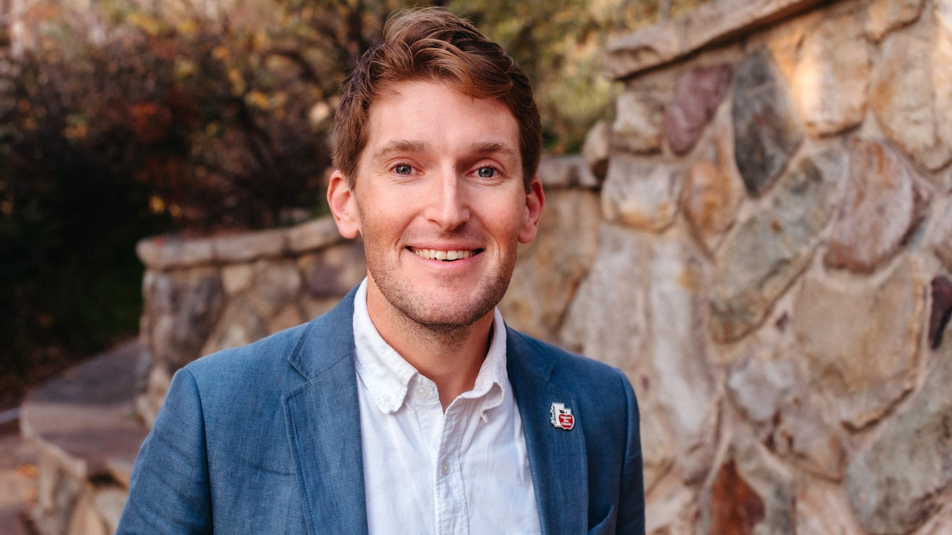 Smiling man in a blue blazer and white shirt stands outdoors beside a stone wall, with autumn trees in the background; a small badge sits on his lapel.