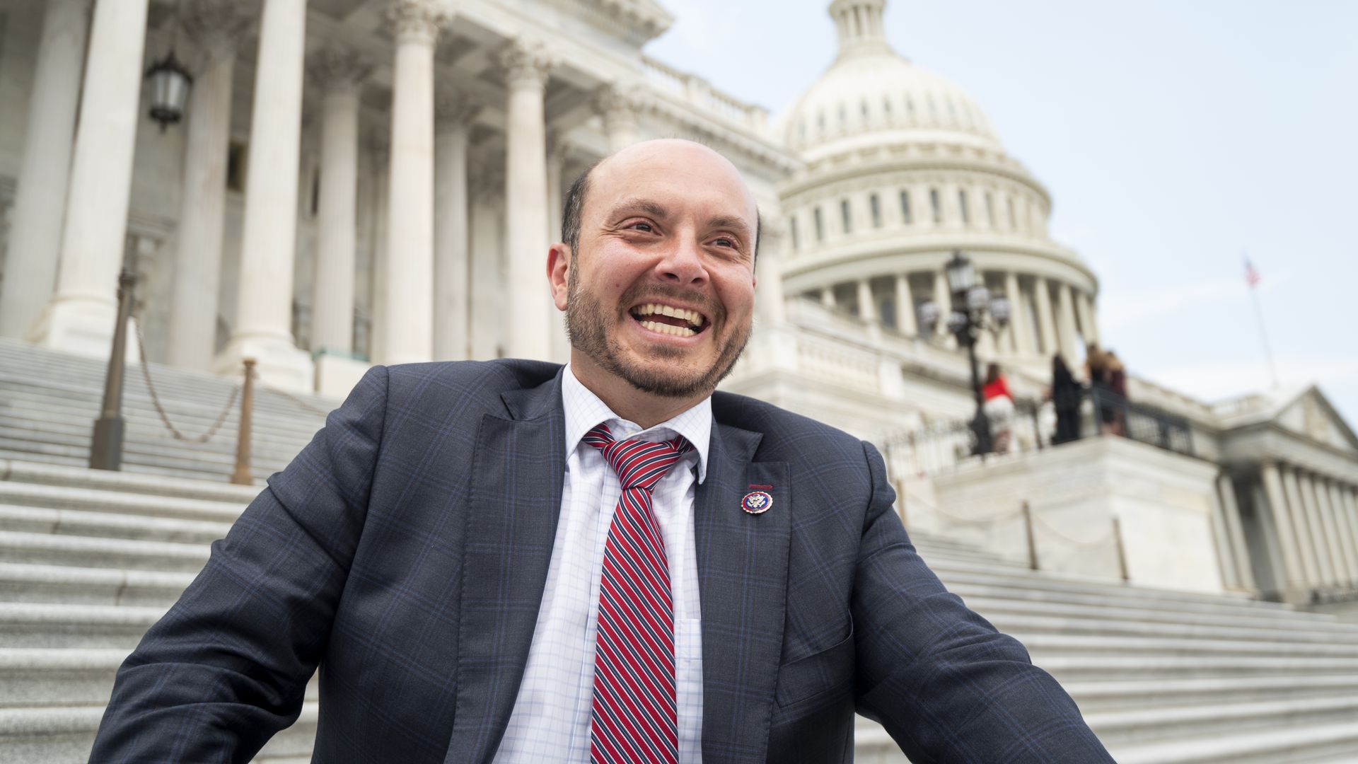 Rep. Andrew Garbarino, wearing a gray suit with a blue grid pattern, a white shirt and a red and blue striped tie, smiles as he sits on the Capitol steps.