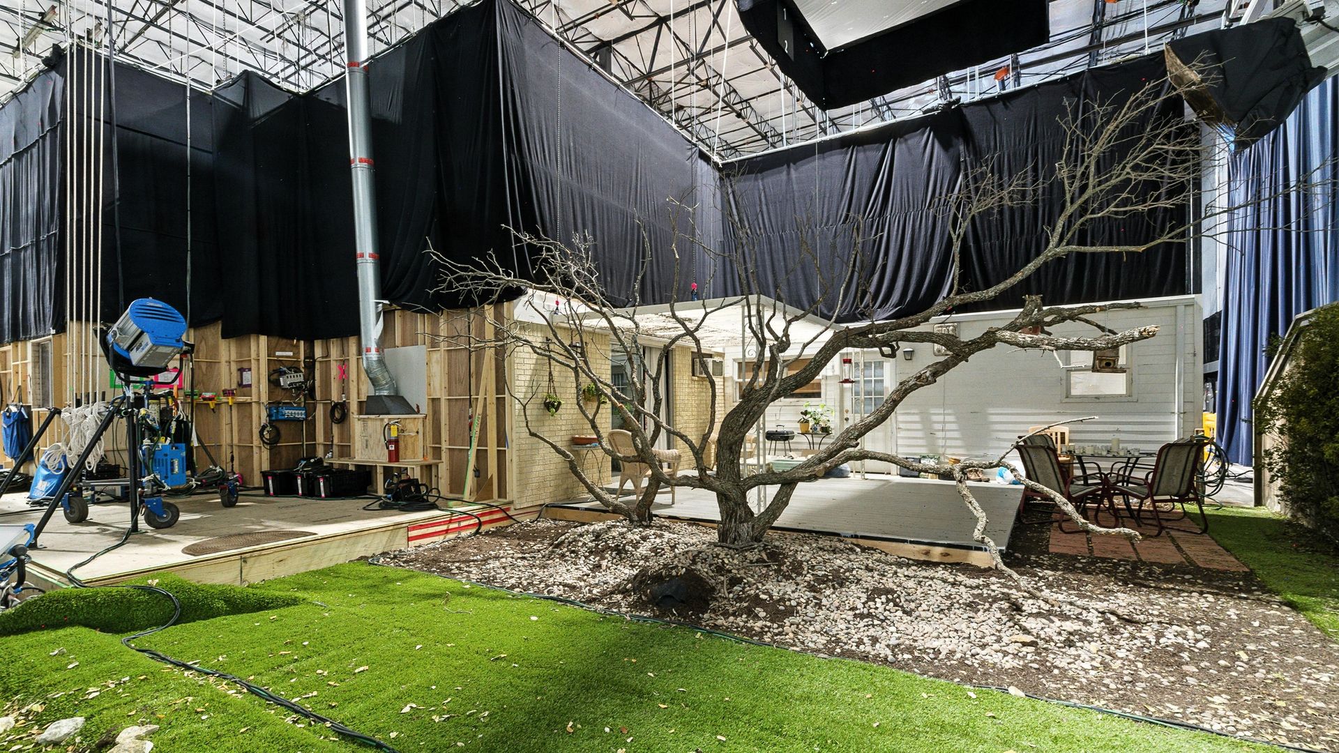 Indoor film set showing a backyard with fake grass, a leafless tree, patio furniture, and a small house facade surrounded by black curtains and studio equipment.