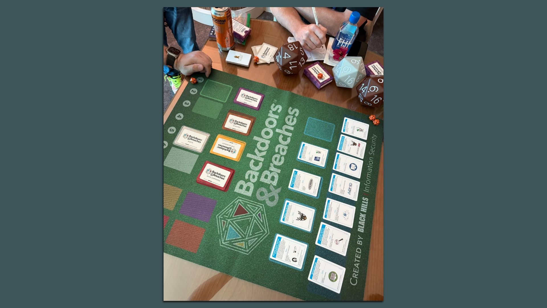 People playing the board game "Backdoors & Breaches" on a wooden table with large dice, cards, and a green play mat showing various colored card decks and game areas.